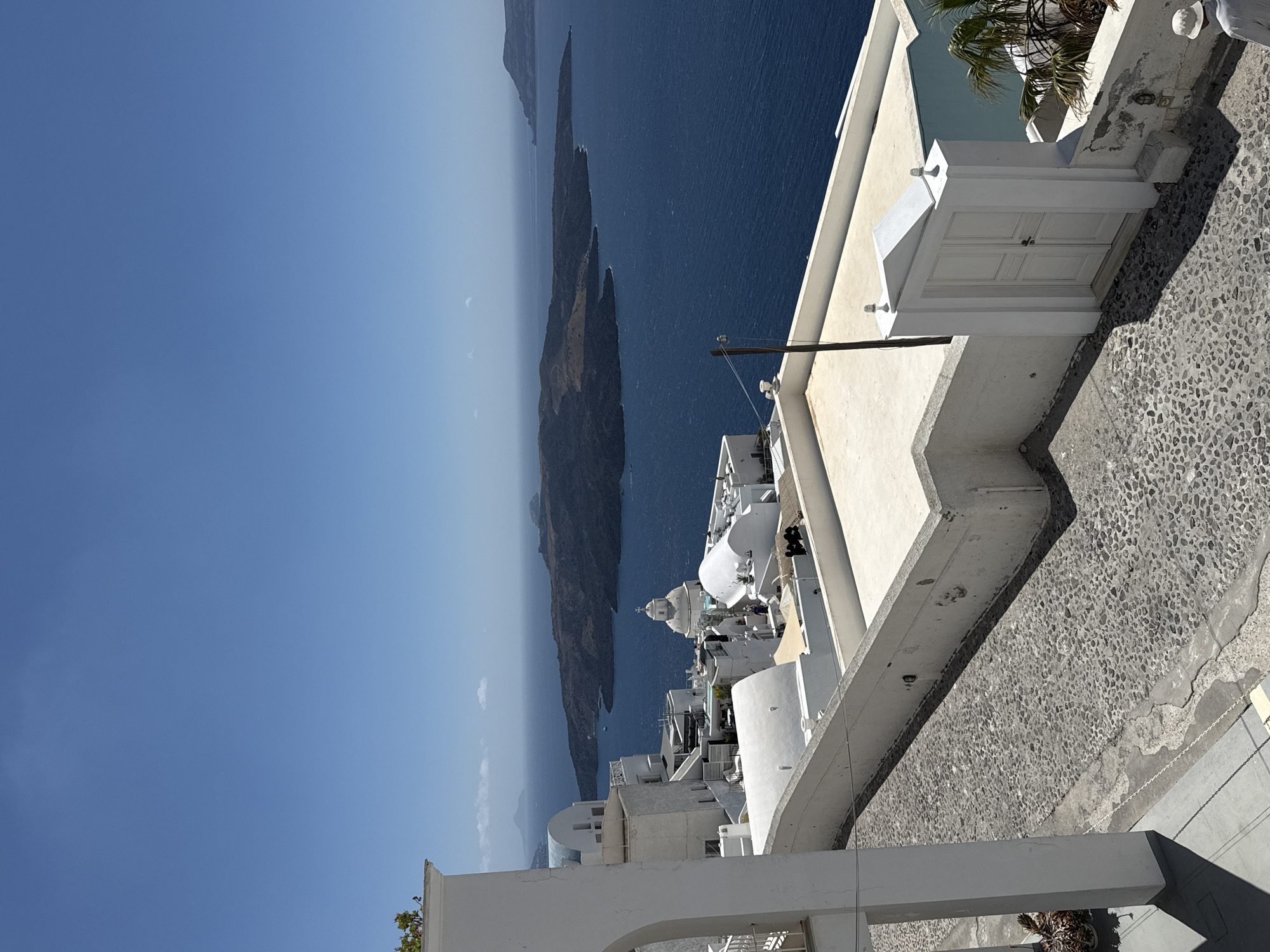 The image depicts a view of the caldera in Santorini, Greece, specifically from the town of Fira. In the foreground, a cobblestone path leads past whitewashed buildings and a white double door. Beyond the buildings, the deep blue waters of the Aegean Sea stretch out, with the volcanic islands of Nea Kameni and Palea Kameni visible in the distance. The sky is clear and bright blue. A few small boats can be seen on the water. The architecture is characteristic of the Cycladic islands, with cubic white buildings, domed roofs, and arched doorways. The overall scene conveys a sense of tranquility and the iconic beauty of Santorini.