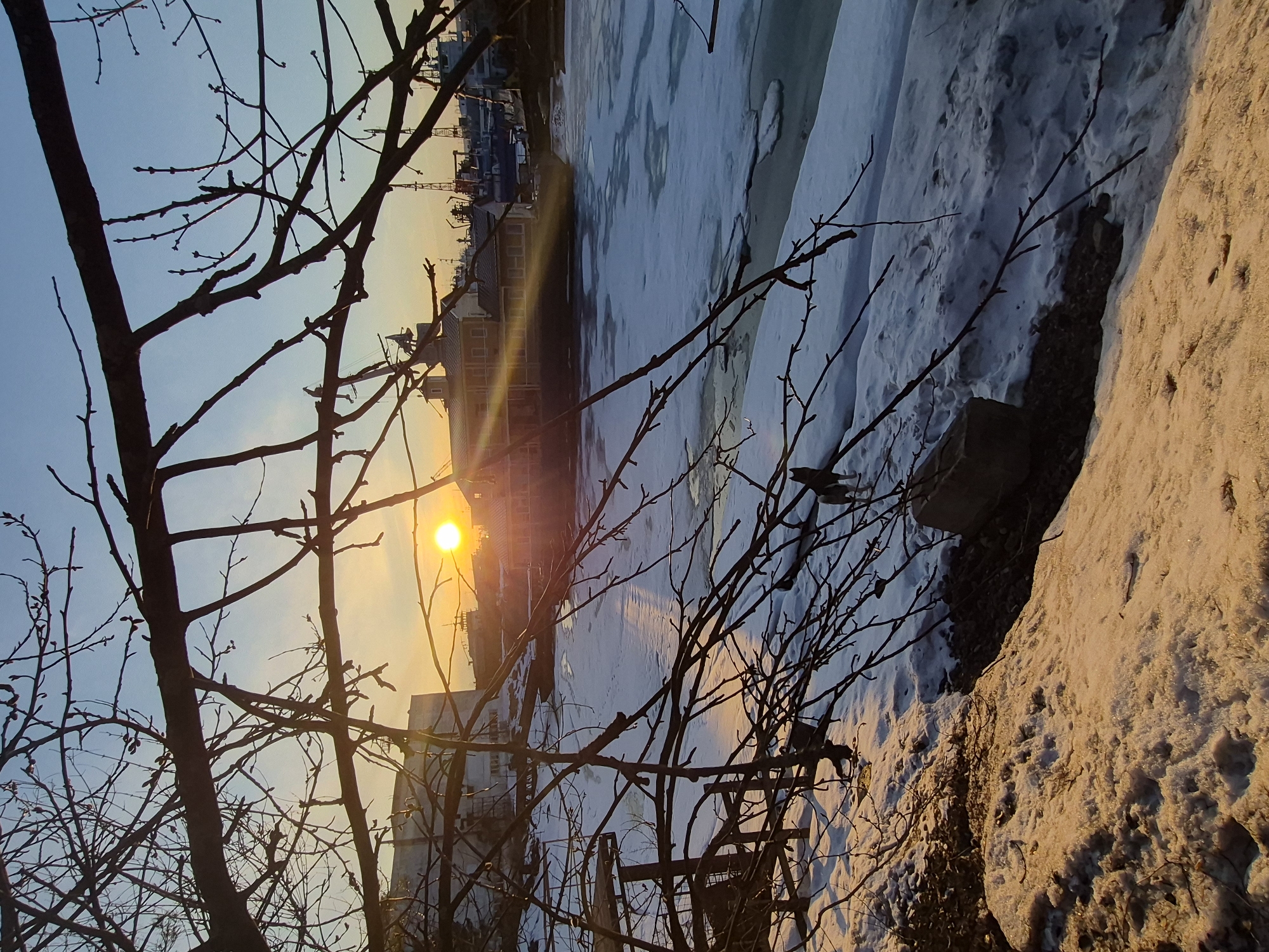 The image shows a winter scene at sunset. The location is Unknown, Unknown. In the foreground are bare tree branches. The sun is visible behind a building, casting a golden glow. A partially frozen body of water is present in the middle ground. The banks are covered in snow. Cranes and docks are barely visible in the distance.