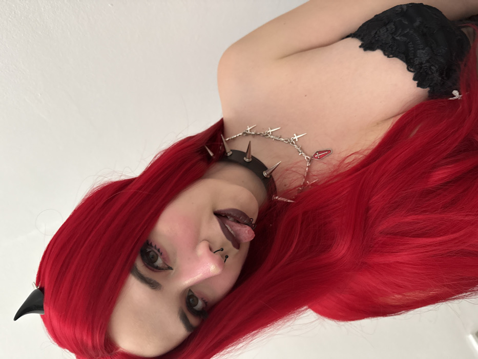 A woman with red hair, styled in voluminous waves, poses with her tongue slightly out. She wears a black choker with spikes and a dangling silver chain adorned with crosses and a coffin charm. Small black horns are visible in her hair. The woman has fair skin, dark eye makeup, and a piercing on her nose. She wears black lace lingerie on her top.