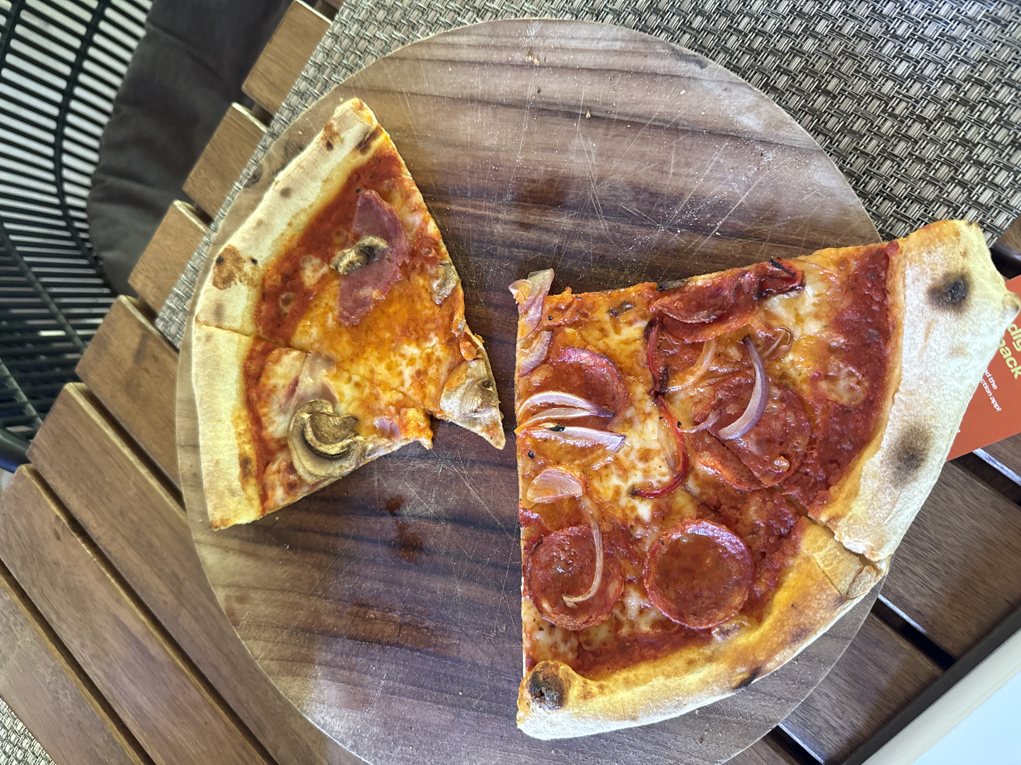 Two slices of pizza are presented on a wooden cutting board, which rests on a woven placemat atop a slatted wooden surface. The pizza on the left appears to have mushroom and pepperoni toppings, while the slice on the right features pepperoni and sliced red onions. A small piece of orange paper with visible text is partially tucked under the right pizza slice. The text on the paper appears to be a brand name or promotional material. The lighting suggests it is daytime. The overall scene depicts a casual meal setting, likely outdoors or in a well-lit indoor space. The pizza crusts are golden brown and appear to be of a thin, crispy style. The toppings are well-distributed on both slices. The wooden board has visible scratches and some scattered flour, indicating previous use. The background shows a slatted wooden structure, possibly outdoor seating or furniture.