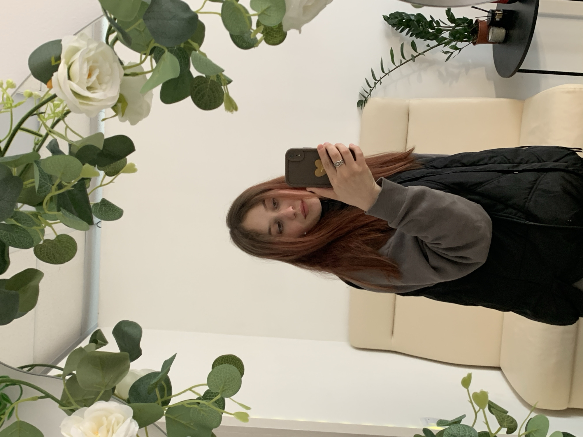 A mirrored selfie shows a young woman with auburn hair in a gray sweatshirt and black vest. She holds a smartphone, taking her own picture. In the mirror's reflection behind her is a cream-colored couch, a black side table, and a potted plant. The mirror itself is decorated with artificial white roses and greenery. The location is consistent with an indoor space, the specific geographic location (Фоки, Russia) seems unlikely to be derived from visual information alone, requiring additional data.