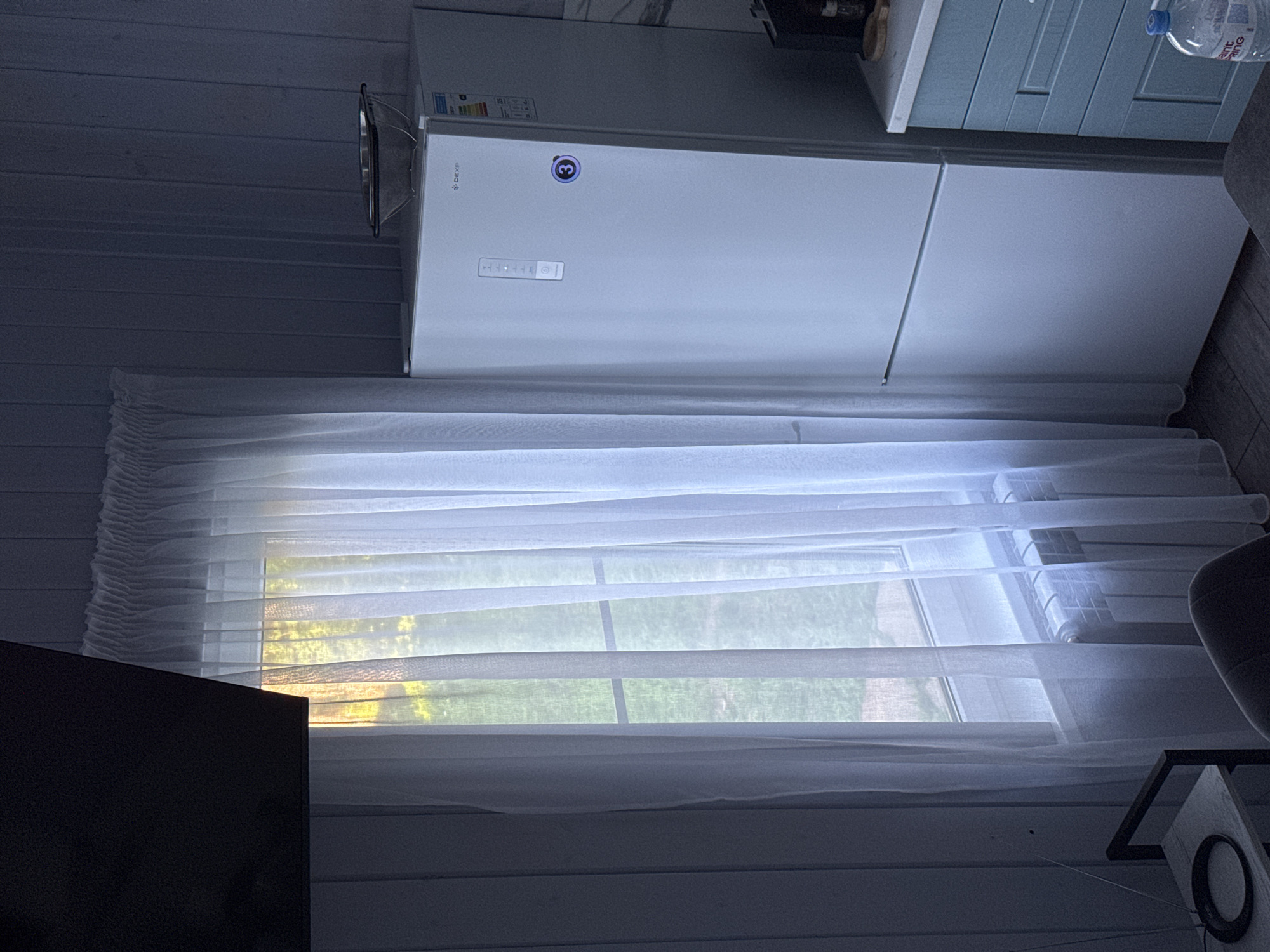 This is an interior shot of a room, likely a kitchen or dining area, in Doma 1096 Km, Russia. A large white refrigerator stands on the right, with a colander on top. Next to it is a blue cabinet with drawers. To the left, a window with sheer white curtains is visible, allowing sunlight to filter through, illuminating the room and showing trees outside. A radiator is situated beneath the window. In the foreground, a dark chair and a small side table with a circular object and an antenna are present. A black object, possibly a television, is partially visible in the top left corner. The walls are paneled.