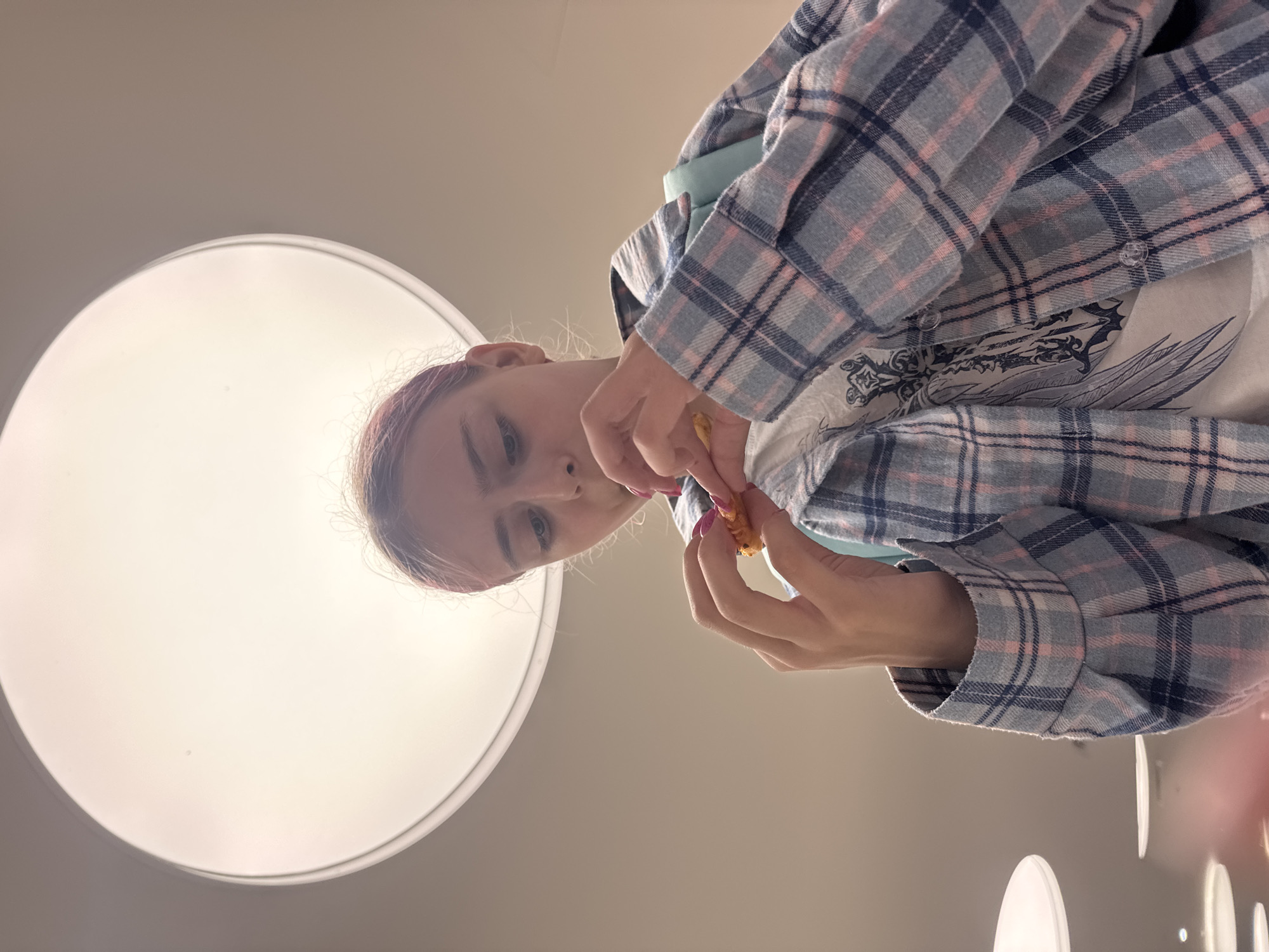 A young woman with pink-tinted hair is seen from a low angle, holding and examining a small, yellow object in her hands. She is wearing a blue and pink plaid shirt over a white t-shirt with a graphic design. Her expression appears focused on the object. In the background, a large, circular, illuminated light fixture is visible against a neutral-colored ceiling. The environment suggests an indoor setting, possibly a public space like a mall or an office building, but no specific Moscow location can be determined.