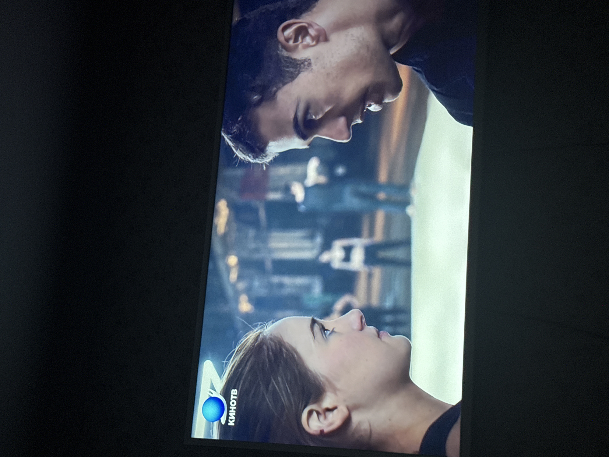 The image shows a screen displaying a scene with two individuals. A woman with light skin and brown hair is looking upwards. Above her, a man with dark hair is looking downwards. The background appears to be an urban setting, possibly indoors. A logo in the lower left corner includes the word "КИНОТВ" (KINOTV) in Cyrillic. The overall setting suggests a romantic movie scene. The location is Mytishchi, Russia based on the Cyrillic writing.