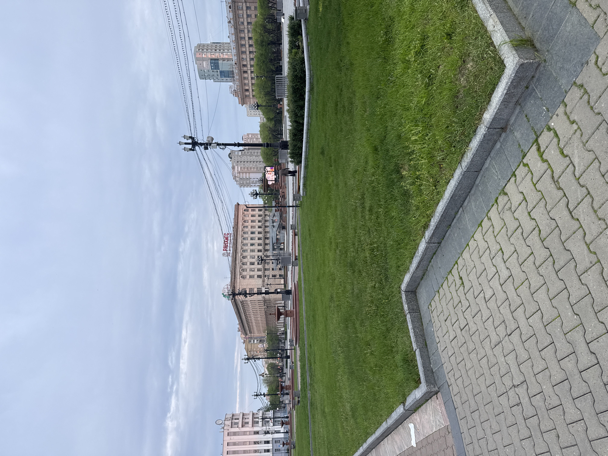 The image depicts a cityscape in Khabarovsk, Russia, featuring a view of the Regional Academy of Public Administration (РАНХиГС) building. The foreground includes a paved sidewalk with a gray brick pattern, adjacent to a grassy area bordered by a concrete edge. The РАНХиГС building is a multi-story structure with a classic architectural design. Above the building, power lines stretch across the sky, which is covered with clouds. Other buildings and urban structures are visible in the distance, with street lamps and a fountain lining the avenue.