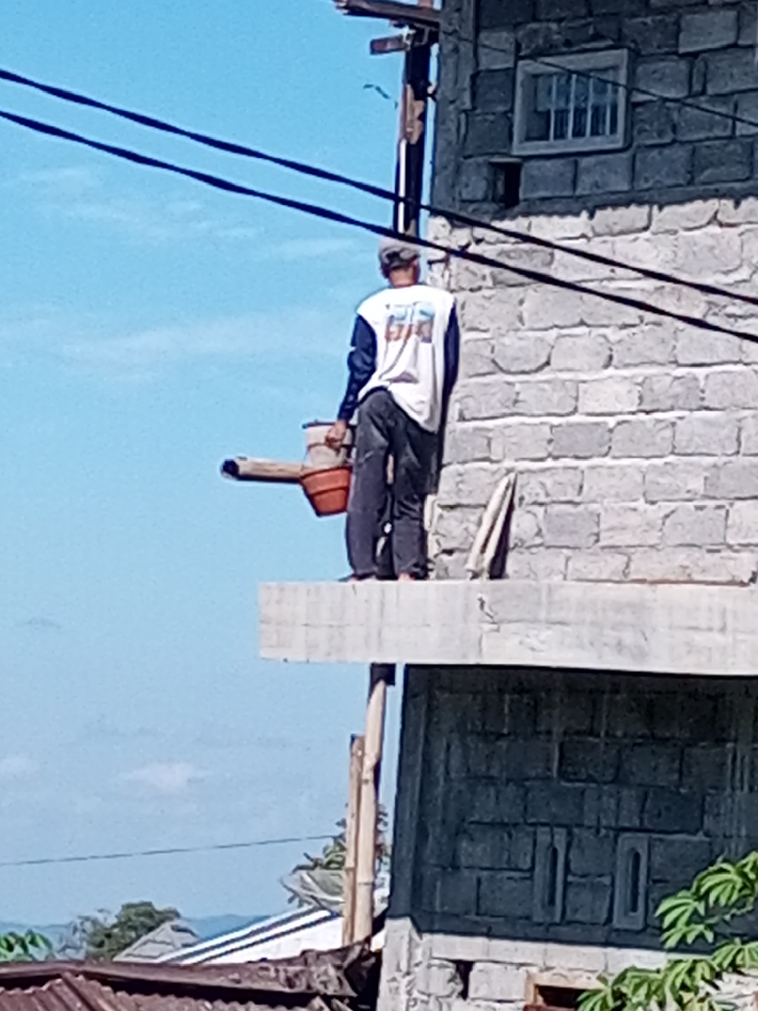 A man wearing a cap, white shirt with blue sleeves, and dark pants stands on a concrete ledge of a brick building, holding a pipe with a red bucket attached to one end. He is outdoors, with a blue sky in the background and power lines crossing the frame. The building has a window with visible panes on the upper floor. In the foreground, parts of a corrugated roof and foliage are visible. The scene appears to be in Andongsili, Indonesia.