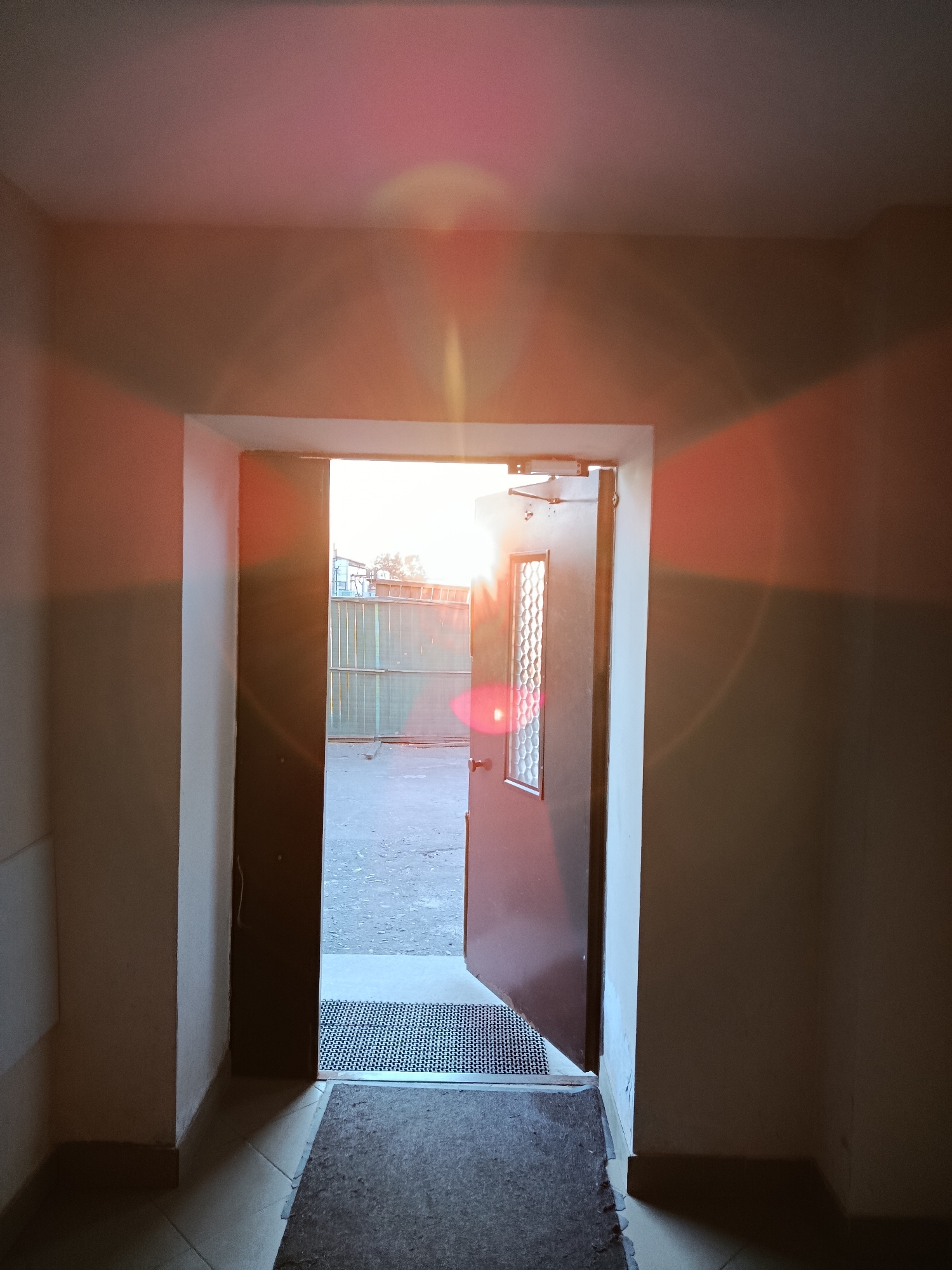 The image depicts the interior view looking towards an open doorway, with the bright light of sunset or sunrise filtering through. The setting appears to be a building's entryway or hallway in Svietlahorsk, Belarus. The walls are painted in a neutral color, possibly beige or off-white, with a distinct shadow cast on the right wall from the doorway. The doorway itself is dark brown metal with a small window pane featuring a textured or patterned glass. A door closer is visible at the top of the door. Inside the doorway, on the floor, are two doormats; a smaller, darker one with a geometric pattern and a larger, coarser one in front of it. Beyond the doorway, an outdoor area is visible, with a corrugated metal fence and what appears to be pavement or a rough ground surface. The sunlight creates lens flare effects, appearing as circular and linear streaks of pink and orange light across the image, particularly on the ceiling and walls. No people or specific actions are discernible in the scene.