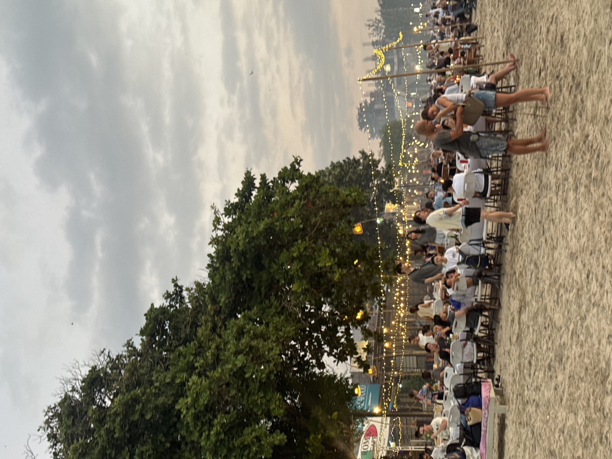 A long shot captures a lively outdoor dining scene on a sandy beach in Kuta Selatan, Indonesia. Numerous tables are set up on the sand, each adorned with white tablecloths and surrounded by chairs. Fairy lights are strung between poles and trees, casting a warm glow over the area. People are seated at the tables, engaged in conversation and enjoying their meals, while others stand and mingle. In the foreground, a couple stands facing each other, seemingly in an embrace or deep conversation. The background reveals more tables and chairs, as well as trees and structures, suggesting a larger gathering or resort area. The sky is overcast, with a few birds visible. The overall atmosphere is one of social gathering and relaxation.