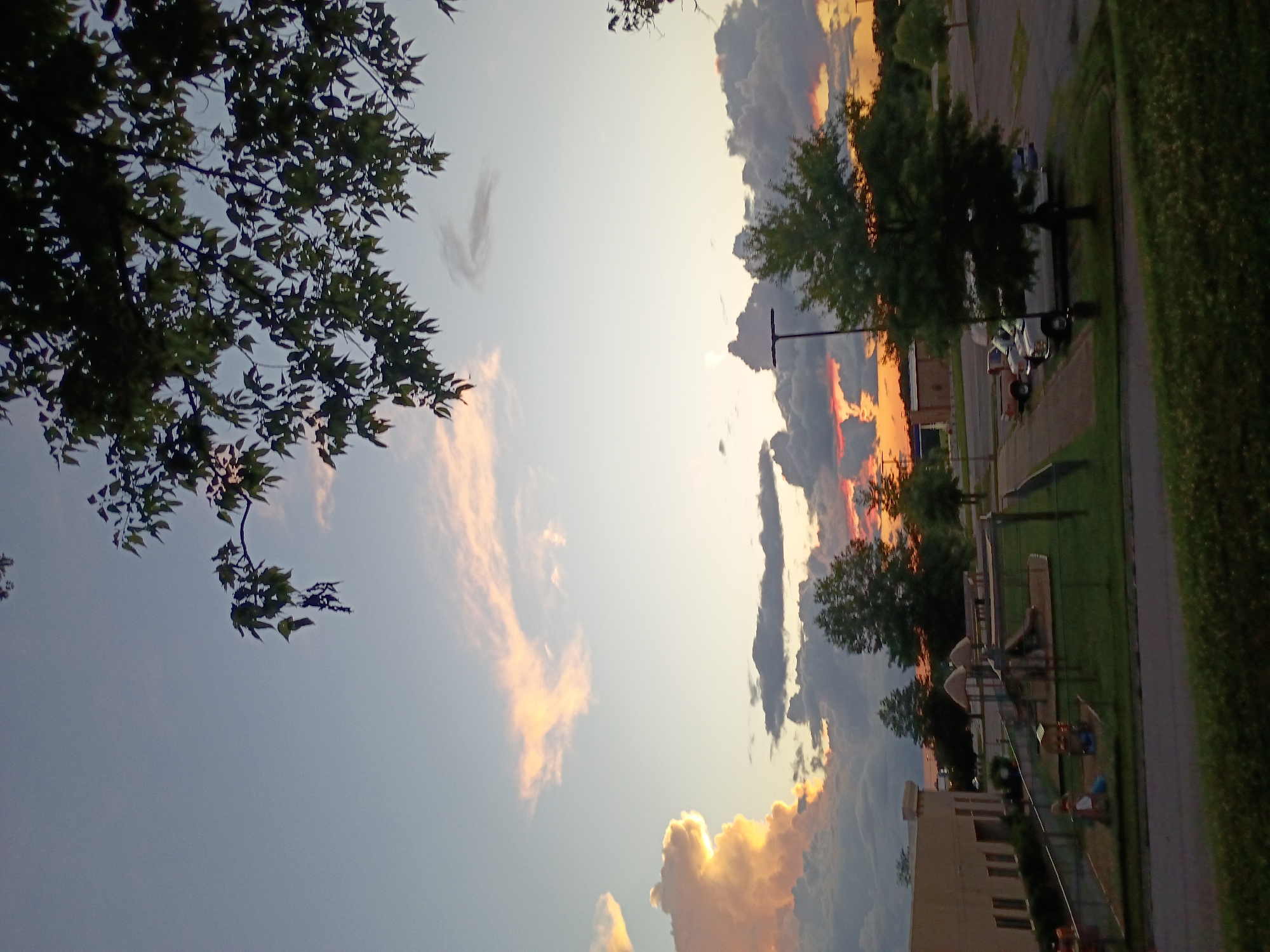The image depicts a twilight scene in Nicholasville, Kentucky, United States of America. A playground with swings and slides is visible, enclosed by a black chain-link fence. To the left of the playground, a building with beige siding and numerous windows stands. Several vehicles are parked in a lot to the right of the playground, including a white van, a pick-up truck, and a light-colored sedan. The sky is filled with dramatic clouds, some illuminated by the setting sun with hues of orange and pink, while others are dark and grey. Trees and foliage are scattered throughout the landscape. The overall impression is that of a quiet, end-of-day scene in a suburban or semi-rural setting.
