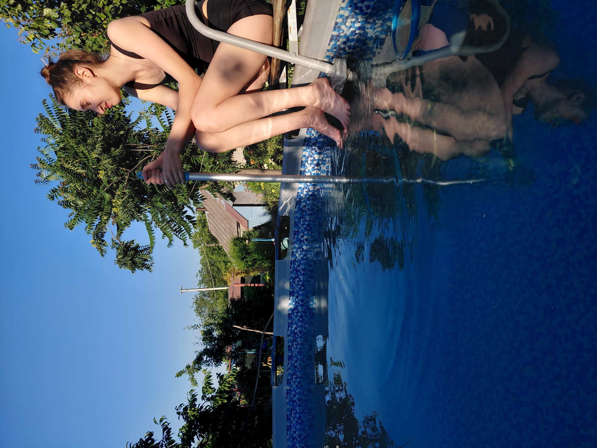 A young woman with brown hair pulled up in a bun sits on the edge of an above-ground swimming pool with her feet submerged in the clear blue water. She wears a black tank top and black shorts. The pool is decorated with a mosaic of blue and white tiles around its rim. A metal ladder is partially submerged in the water on the right side of the pool. Sunlight casts shadows on the water, creating patterns of light and dark. The background shows a residential area with houses, trees, and a clear blue sky. The woman is holding a silver pole in her hands.