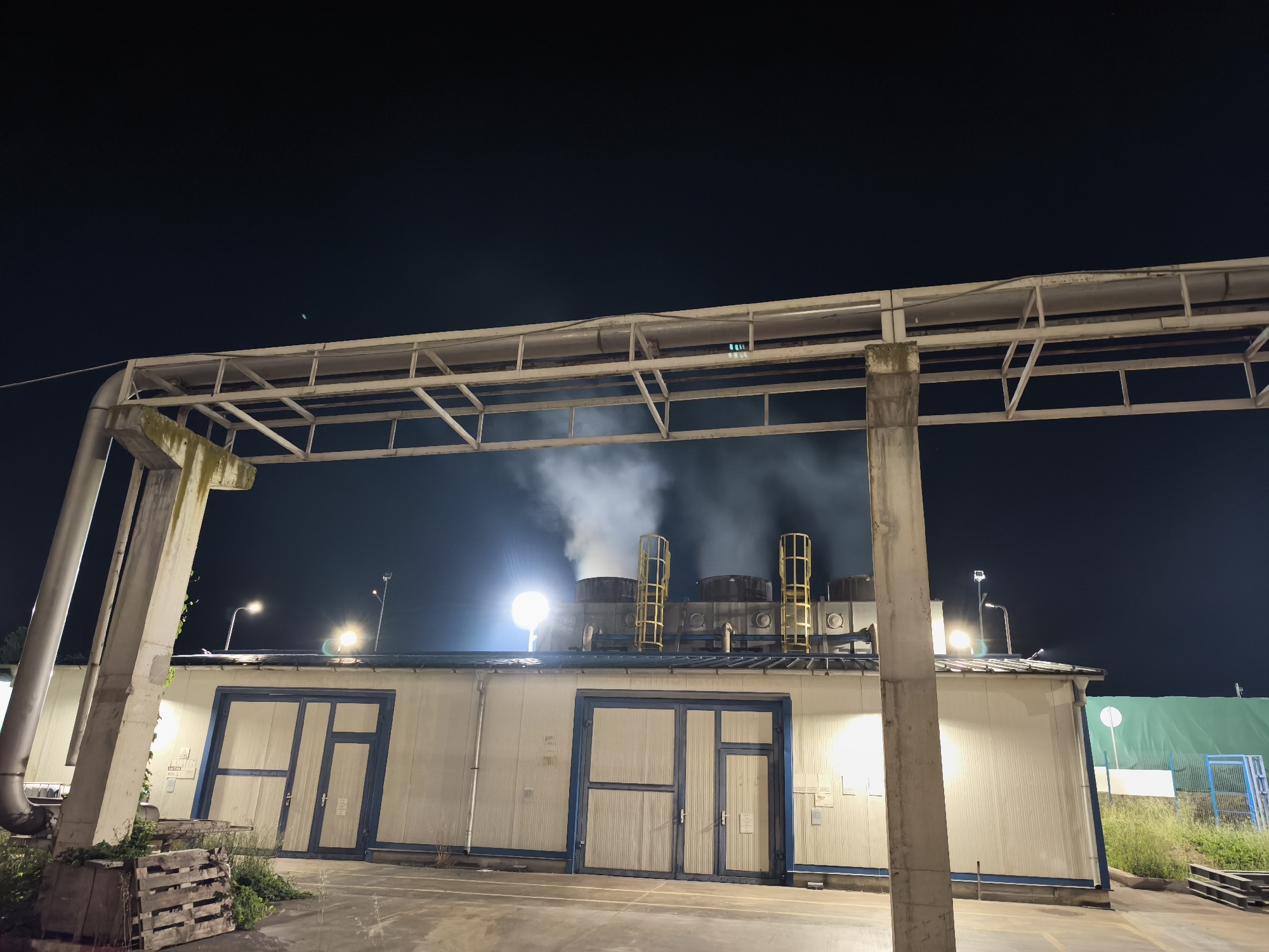 The image depicts an industrial scene at night. In the foreground, a large, white, rectangular building with blue trim and large double doors is visible. To the left, a concrete pillar supports a metal beam structure that extends across the top of the frame. In the background, white smoke billows from industrial equipment on the roof of another structure. The scene is illuminated by artificial lights, casting a glow on the building and the smoke. The environment appears to be an outdoor industrial area, with some greenery and wooden pallets visible.