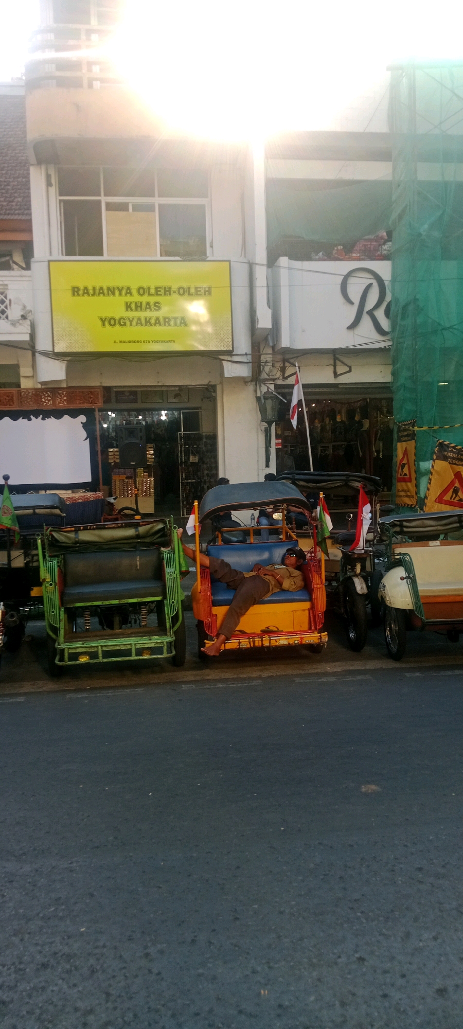 The image depicts a street scene in Yogyakarta, Indonesia, with a focus on a row of parked vehicles, likely used for transportation or tourism. In the foreground, a man is asleep, reclined in the passenger seat of a yellow and blue auto-rickshaw. He is wearing sunglasses, a light-colored shirt, and dark pants. His left leg is propped up and extended. To the left of this auto-rickshaw is a green and black auto-rickshaw. Further to the right, another white and cream-colored vehicle is partially visible, possibly another auto-rickshaw or a small van.
In the background, a building with a yellow sign is prominently displayed. The sign reads "RAJANYA OLEH-OLEH KHAS YOGYAKARTA" in large text, indicating it is a shop selling special souvenirs from Yogyakarta. Below this, smaller text reads "JL. MALIOBORO 67A YOGYAKARTA," identifying the specific location on Malioboro Street. The building has a storefront with an open entrance, revealing goods displayed inside. Above the shop, the building's facade shows windows and architectural details. To the right of the shop, a large black "R" is visible on the building's exterior, suggesting a brand name. Construction netting covers a portion of the building on the far right. Indonesian flags are visible, one attached to a pole near the shop entrance and smaller flags on the auto-rickshaws. The scene appears to be a public area, possibly a tourist hub.