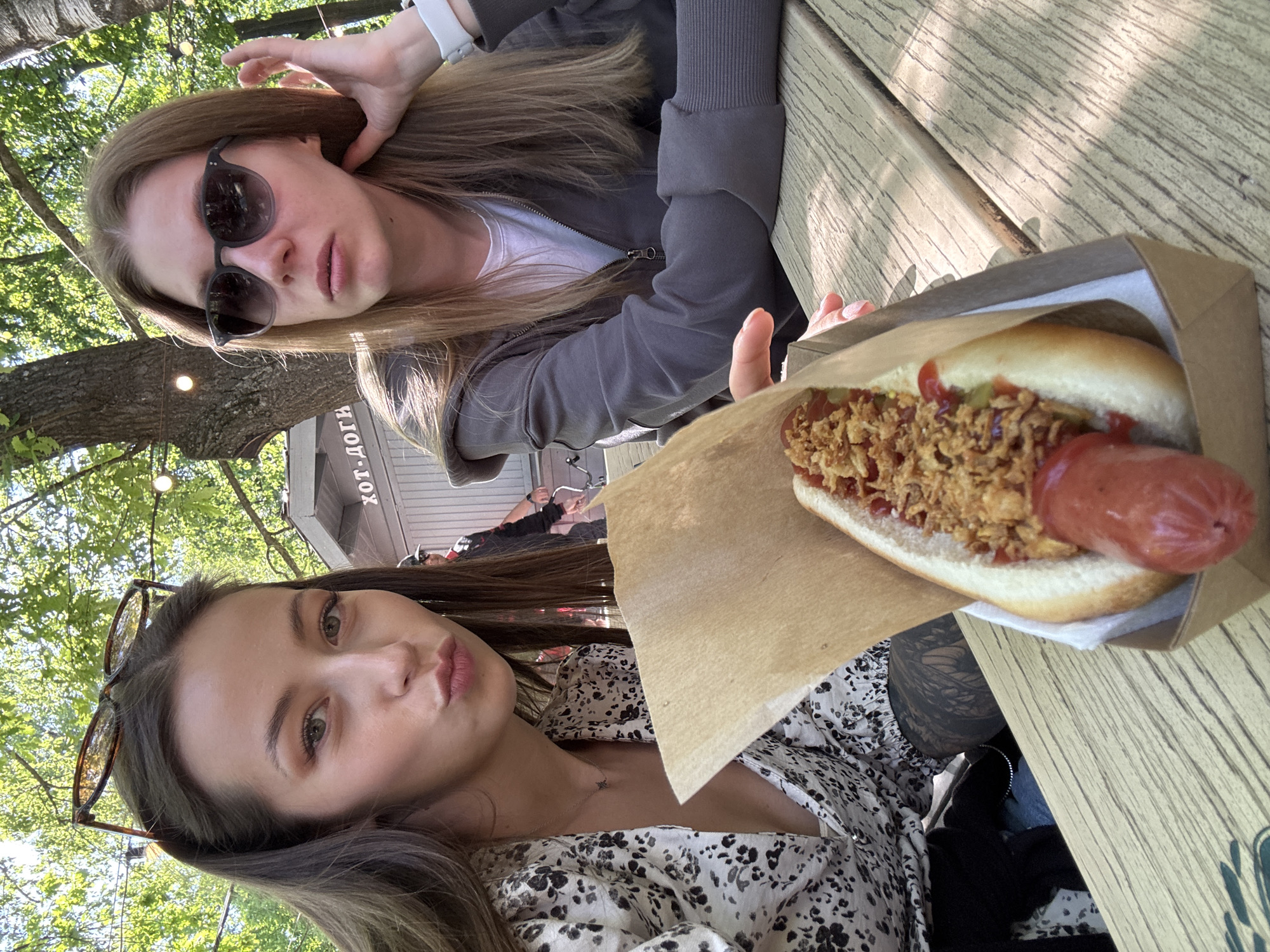 This outdoor selfie captures two young women at a casual eatery in Saint Petersburg, Russia. They are seated at a light-colored wooden picnic table, with lush green foliage and string lights visible in the background, indicating a daytime setting.
In the upper part of the image, one woman with long, light brown hair wears sunglasses and a gray zip-up hoodie over a white top. A white wristband is visible on her left wrist, and she holds her left hand near her head in a gesture.
Below her, another woman with long, dark brown hair and a white and black patterned top looks towards the camera with slightly pouted lips. A pair of sunglasses rests on top of her head, and she wears a delicate necklace.
Between the two women, a hot dog in a brown paper tray is prominently displayed. The hot dog features a large sausage, topped with crispy fried onions, ketchup, and what appears to be mustard or relish.
In the background, behind the table and beneath the trees, part of a building or stand is visible. A sign on this structure clearly displays the Cyrillic text "Хот-доги" (Khot-dogi), which translates to "Hot-dogs" in Russian, confirming the establishment's offerings. The bright sunlight creates dappled shadows on the table, contributing to the casual outdoor atmosphere. The women appear relaxed, enjoying their meal and posing for the photo.