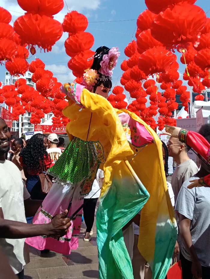 This image captures an outdoor cultural event in Nairobi, Kenya, under a clear blue sky. Hundreds of red, accordion-style Chinese lanterns are suspended overhead, forming a dense canopy across the upper frame. In the central foreground, a large, elaborate traditional Chinese puppet is being operated. The puppet features a pale-colored face, a dark ornate hairstyle adorned with pink and yellow flowers and white pearl-like beads, a yellow upper garment, and a lower garment composed of a green sequined bodice over pink fabric with embroidered patterns. Flowing yellow and green fabric streamers extend from the puppet's sides. A dark-skinned individual, wearing a light-colored t-shirt, is visible on the left, operating the puppet using a dark rod held in their right hand. This individual is partially visible, with their left arm and a portion of their smiling face. The mid-ground is occupied by a diverse crowd of individuals, largely out of focus, dressed in casual clothing. White event tents or stalls with partially visible signage, including Chinese characters, are situated in the background. Modern, multi-story white buildings are visible in the far distance beneath the lanterns. The ground beneath the attendees appears to be paved with large, light-colored square tiles.