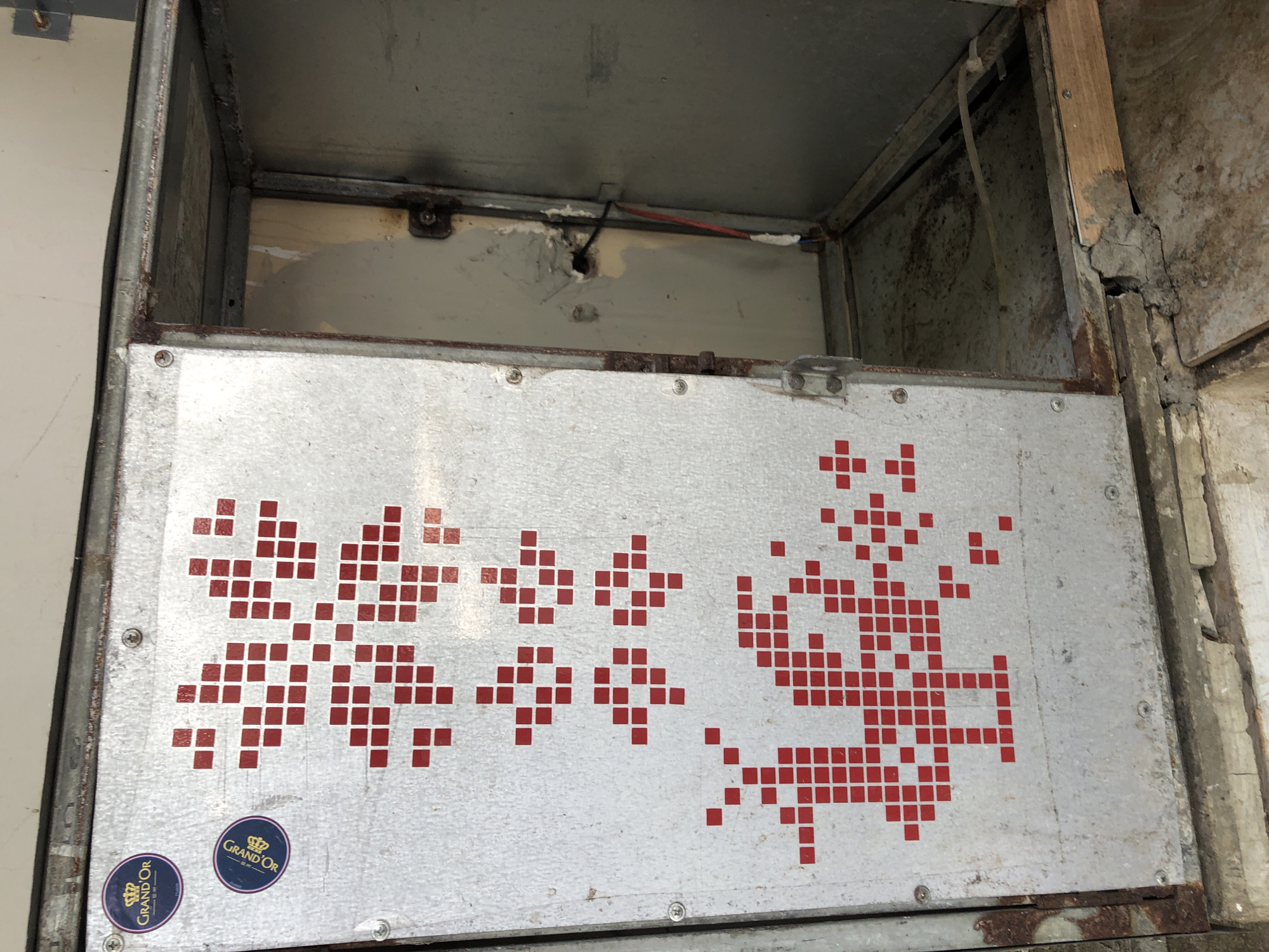 The image depicts an open metal cabinet, possibly an air conditioning unit or ventilation access panel, set against a brick and concrete base. The cabinet is weathered, with visible rust on its metal surfaces and along the edges. On the left panel of the cabinet, there are two circular blue and gold stickers that read "GRAND OR" and a red pixelated design resembling a snowflake or a stylized bird. The interior of the cabinet is visible and appears empty, with wires and some debris present. The cabinet is located on the exterior of a building, suggesting it is an outdoor unit. The scene is observed from a slightly elevated perspective, looking downwards into the open cabinet. The environment appears industrial or utilitarian, with the worn surfaces and exposed components indicating a functional rather than decorative purpose. There is no indication of the specific location in Da Nang, Vietnam, beyond the general context of an outdoor unit.