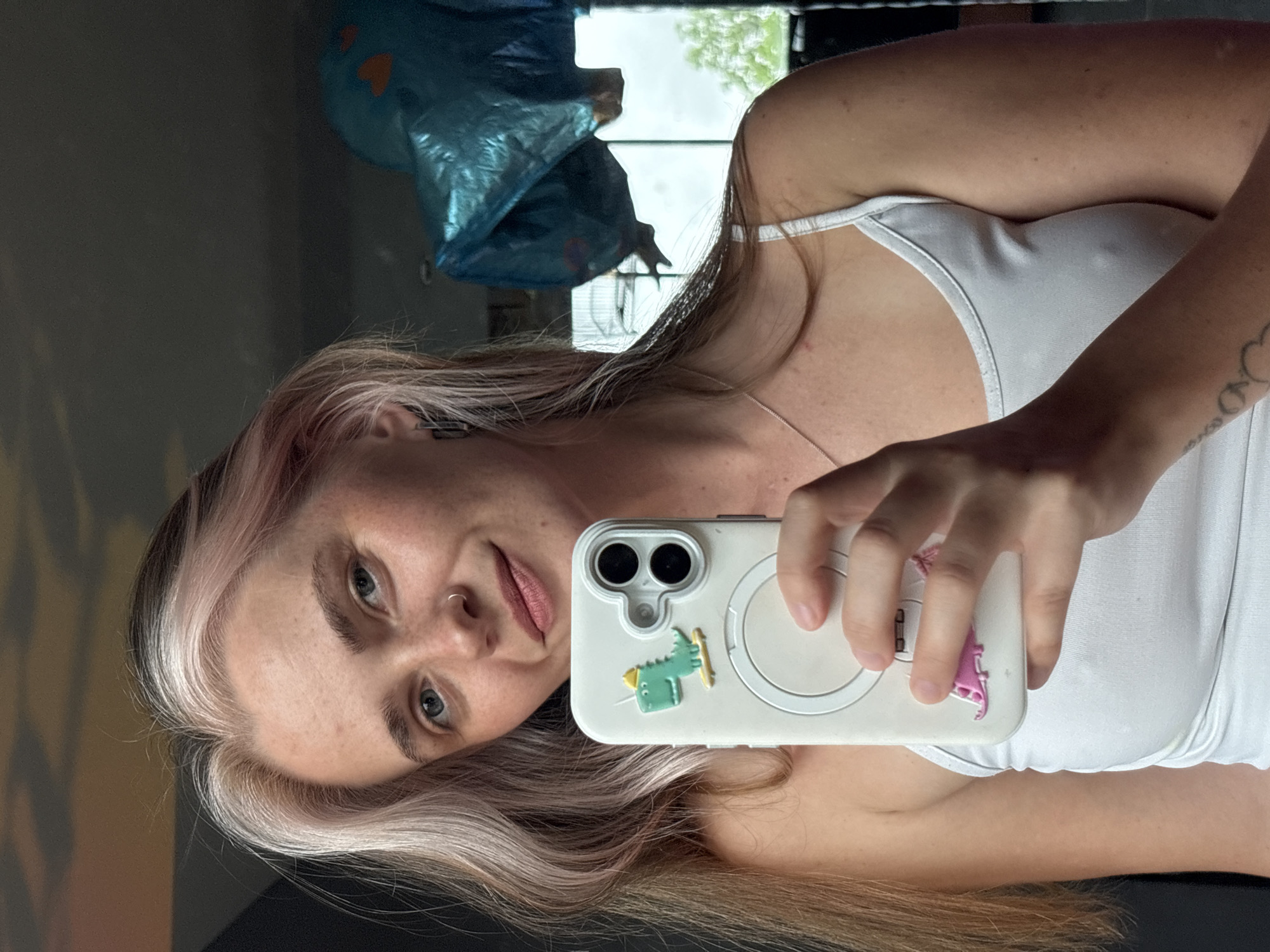A young woman with blonde and pink streaked hair and blue eyes takes a selfie in her bedroom. She is wearing a white tank top and has a silver necklace on. Her phone is decorated with two dinosaur stickers. In the background, a large blue inflatable object is visible, as well as a window showing greenery and a cloudy sky.
