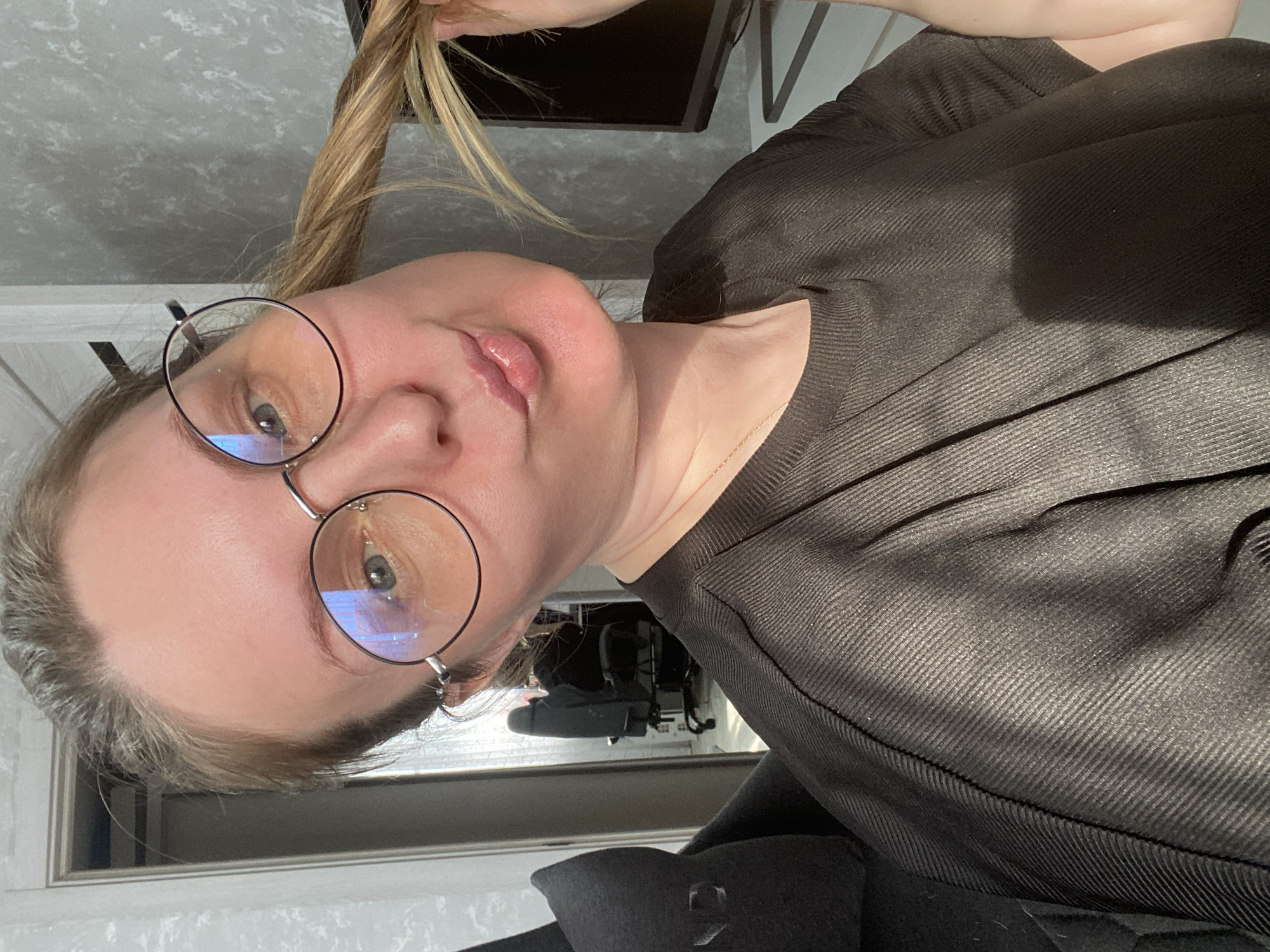 This close-up portrait shows a person, likely female, indoors in Noviy, Russia. The individual has fair skin and light brown hair with blonde highlights, which is held by a hand near the top of their head. They are wearing distinctive round, wire-rimmed glasses, in which blue light reflections are visible. The person gazes directly towards the viewer with a neutral expression, their lips slightly parted in a soft pout.
They are dressed in a dark, possibly black or dark brown, top featuring a prominent ribbed or textured pattern running diagonally. A thin chain necklace is visible around their neck. A hint of another dark garment, potentially a coat or jacket, can be seen in the lower right portion of the image.
The background is softly out of focus, revealing an indoor setting. A dark, rectangular object, possibly a monitor or television screen, is partially visible behind the person's head. The lighting is bright and appears to be natural, suggesting it is daytime, with light casting subtle shadows on the person's face and clothing. There is no visible text in the media file.