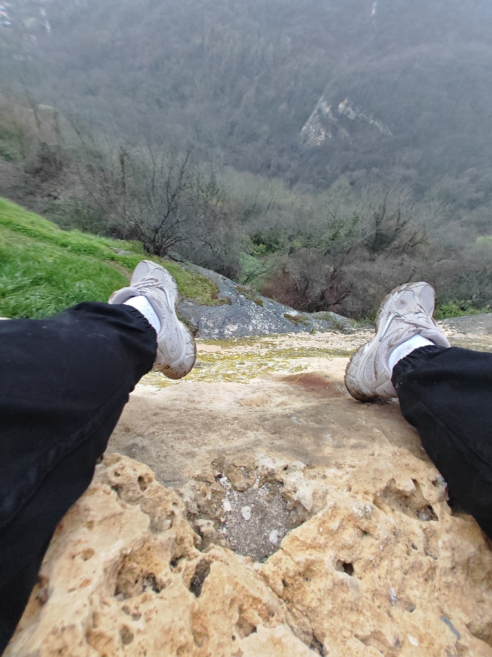 The image captures a first-person perspective looking downward from a high vantage point in the city of Shumen, Bulgaria. In the foreground, the lower legs and feet of an individual are visible, clad in black trousers, white socks, and light grey athletic shoes. The feet are positioned above a textured, light brown rock ledge, suggesting the person is seated or lying down, with their legs extended over the edge. Directly beneath the rock ledge and extending into the midground is a steep incline featuring patches of green grass, dark grey rock, and sparse, bare deciduous trees. The background reveals a deep, forested valley dominated by numerous deciduous trees, primarily devoid of leaves, indicating a non-summer season. The distant terrain shows a mix of dark tree cover and lighter, exposed rock or earth. The sky is overcast, contributing to a muted, diffused light across the entire scene.