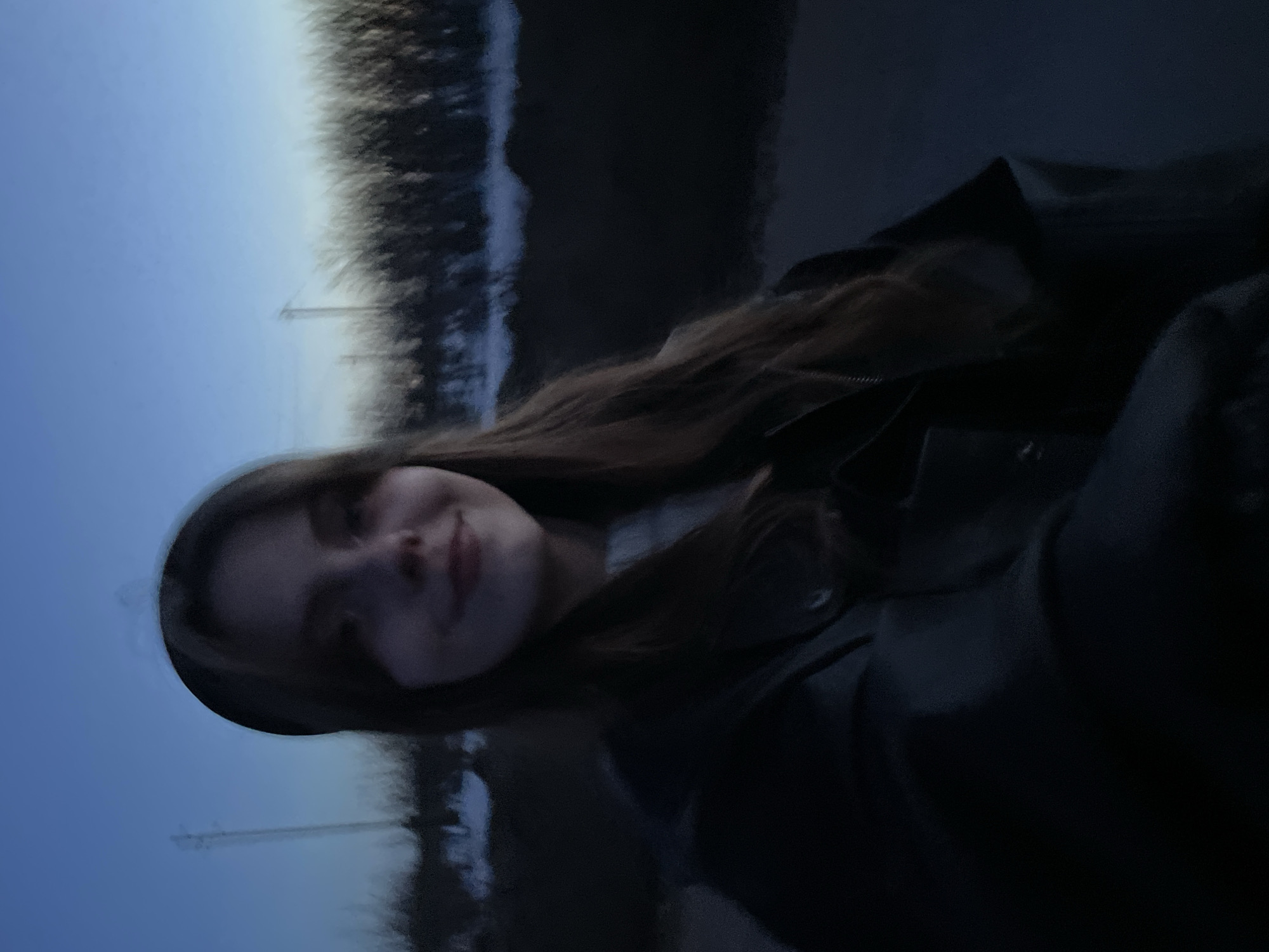 A young woman with long brown hair and fair skin is wearing a dark jacket and smiling slightly. She is positioned in front of what appears to be a body of water reflecting the sky. Telephone poles are visible in the background. The photo seems to have been taken outdoors in low light, potentially during dusk or dawn. The city of Фоки, Russia cannot be determined from the image.