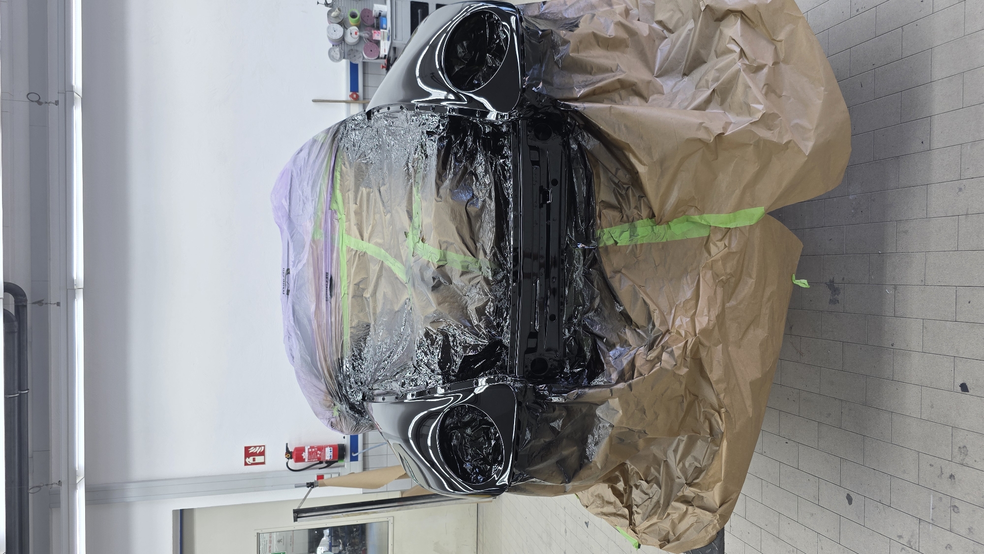 A black car, covered in brown paper and clear plastic sheeting, is parked indoors. The sheeting is secured with green tape. The car's headlights are visible. In the background, a white wall with fluorescent lighting is present. A red fire extinguisher is mounted on the wall to the left. Shelving units with various items are visible in the background to the right. The floor is tiled.