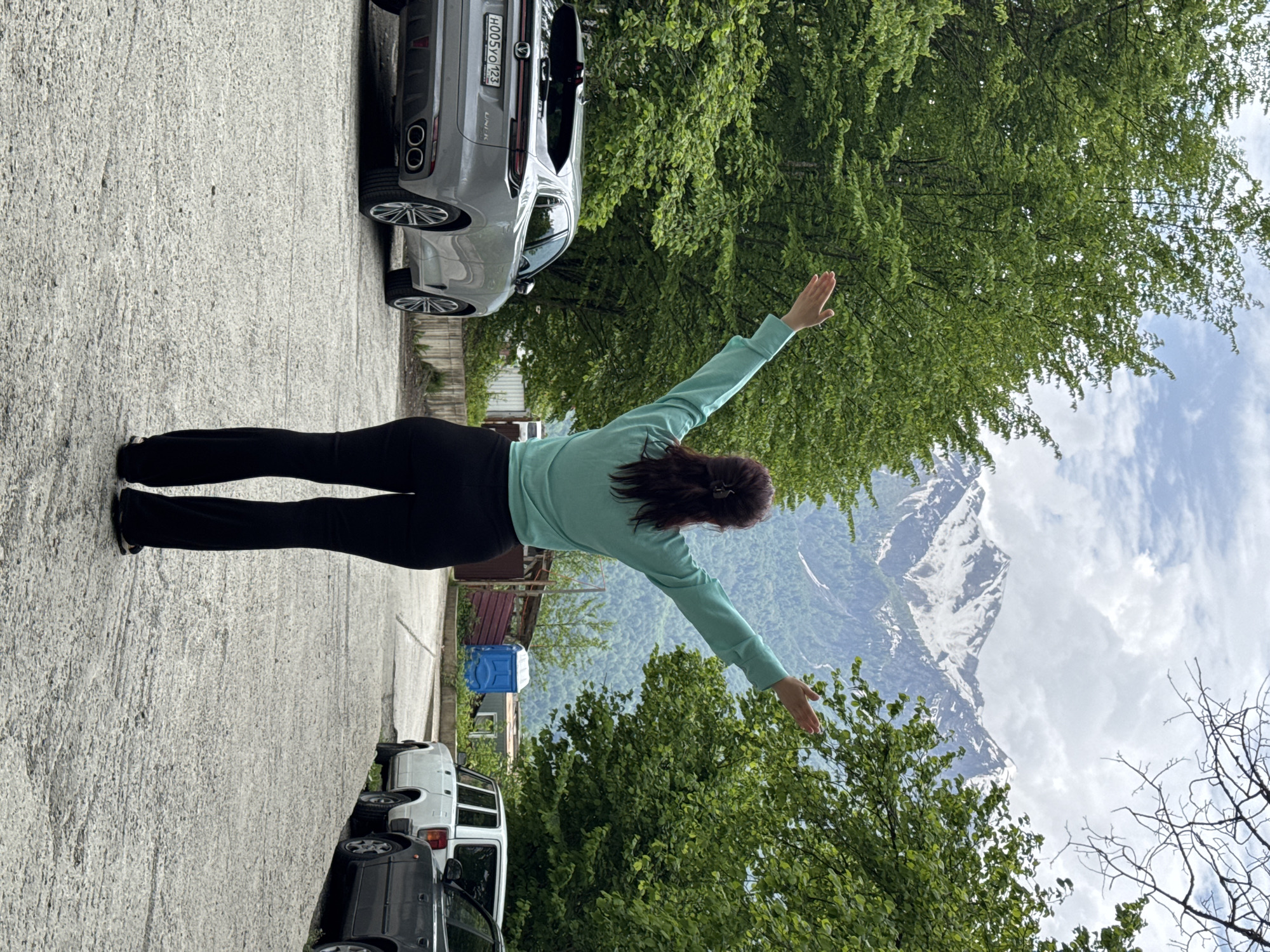 A woman in black leggings and a light-blue sweater stands with her arms outstretched in front of a mountainous landscape in Krasnaya Polyana, Russia. The mountains in the background are partially covered in snow. Trees fill the foreground and midground. The woman stands on a concrete surface, with parked cars on either side. The license plate on the grey car reads "H005YO123."