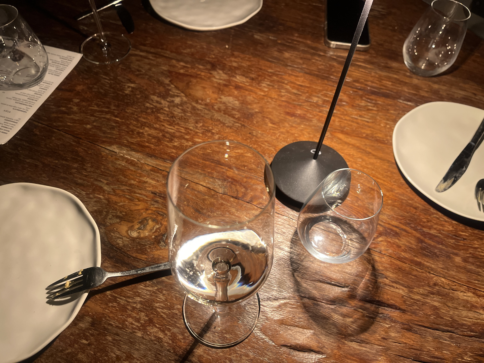 A photograph captures a dining scene on a dark wooden table. In the foreground is a water glass partially filled with water. Beside it is a smaller, empty glass. A fork, slightly used, rests on a white plate to the left. Further back, another white plate is set with a knife and fork. In the background, additional dining items are scattered, including another water glass and a menu. A modern black table lamp with a thin stem is also visible. The scene appears to be in a restaurant setting in Kuta Selatan, Indonesia.