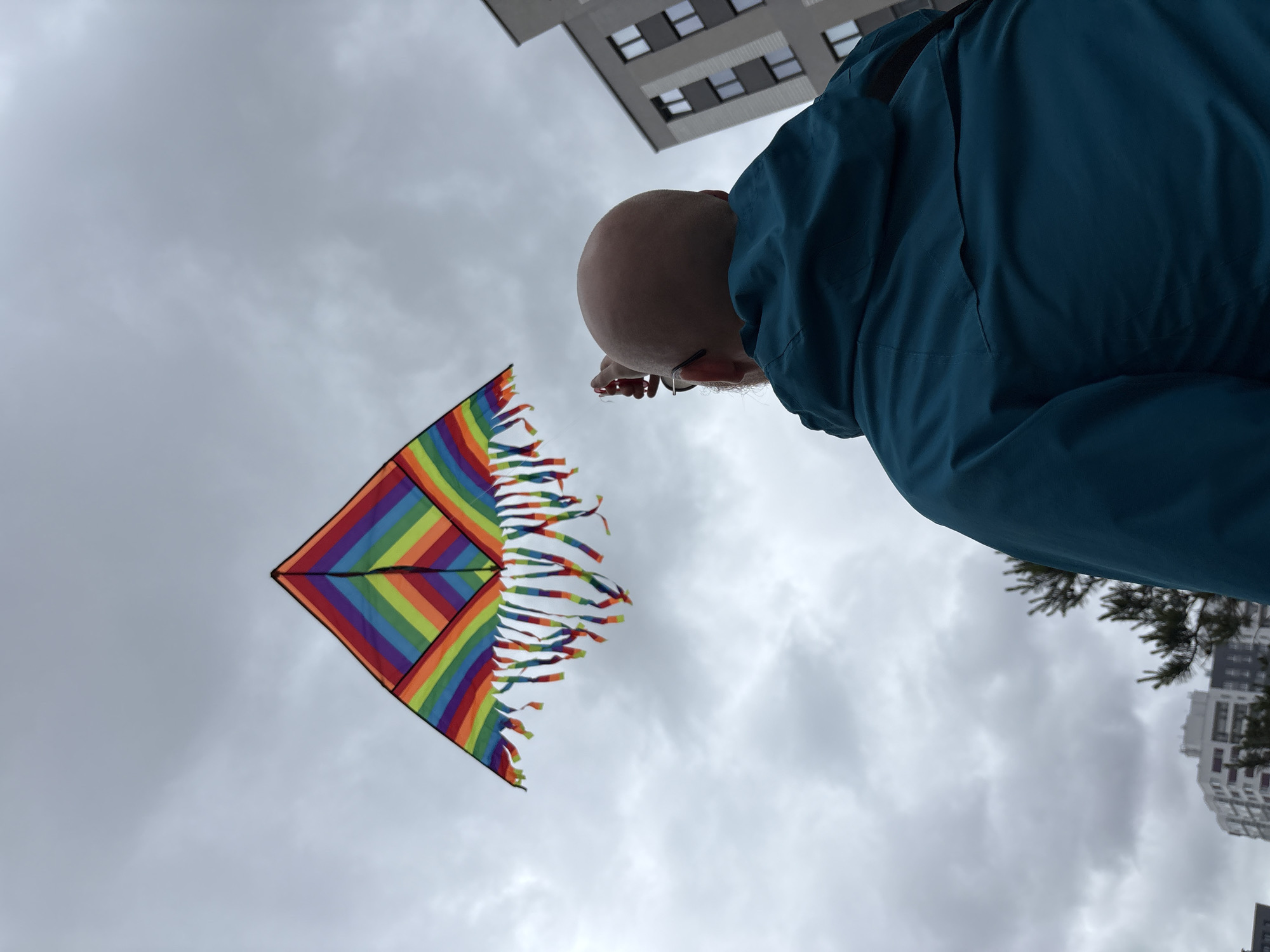 An outdoor, low-angle shot depicts a bald, bearded man in a teal jacket flying a rainbow-colored kite against an overcast sky. The man, seen from behind and slightly to his right, wears dark-rimmed glasses and holds a white kite string reel in his raised right hand, directing his gaze upwards towards the kite. The kite, featuring a diamond-like shape with horizontal stripes of red, orange, yellow, green, blue, and purple arranged in diagonal sections, has numerous colorful fabric fringes along its bottom edge. It is positioned high in the upper-left quadrant of the frame. The sky is largely covered with grey, diffused clouds. In the background, modern multi-story residential buildings are visible; a grey building with dark windows occupies the right side, and lighter-colored structures appear further back on the left. A few dark green evergreen trees are also present in the mid-ground. The specific location within Izhevsk, Russia, is not discernible from the visual evidence.
