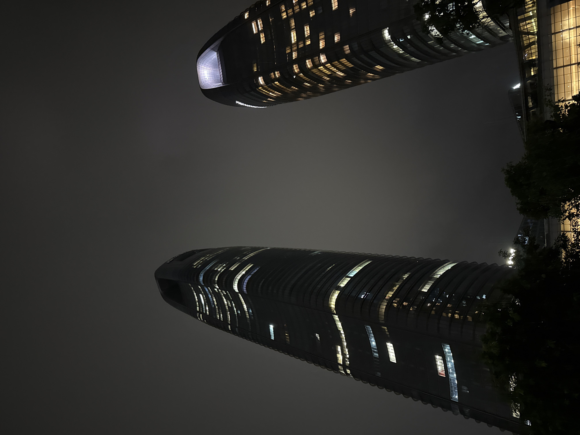 The photograph captures two modern skyscrapers at night in Haizhu District, China. The buildings have sleek, curved designs and are illuminated by numerous lit windows, suggesting occupancy. The sky is dark and overcast, creating a muted and atmospheric backdrop for the illuminated structures. In the foreground, the tops of dark green trees are visible, partially obscuring the lower sections of the buildings and the surrounding environment. The overall impression is one of urban grandeur and nighttime activity.