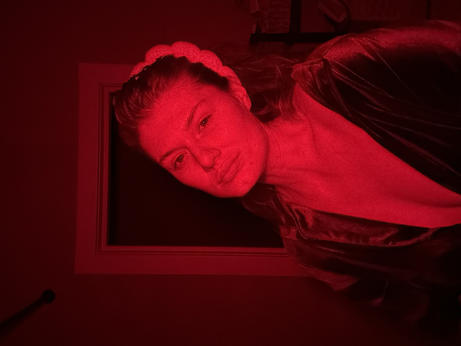A young woman is captured in a medium close-up shot, bathed entirely in a strong, pervasive red light. Her head and upper torso are visible, positioned diagonally across the frame, looking directly towards the viewer. She has light-colored hair, with some strands framing her face, and is wearing a light-colored, possibly cream or white, braided or padded headband. Her face, illuminated in red, shows subtle details, including visible freckles. Her eyes are open and engaging, and her lips are softly closed or slightly parted.
She is wearing a dark, possibly velvet or satin, wrap-around robe or jacket, which reflects the red light with a gentle sheen. The fabric appears soft and luxurious.
The background is dark and indistinct, also saturated with the red hue. Behind her head and shoulder, a rectangular opening, possibly a doorway or window, is visible, outlined by a lighter-colored frame (perhaps white or light wood). The overall impression is one of intimacy and striking mood, created by the dramatic red lighting, which casts deep shadows and highlights contours. No text is visible in the image.