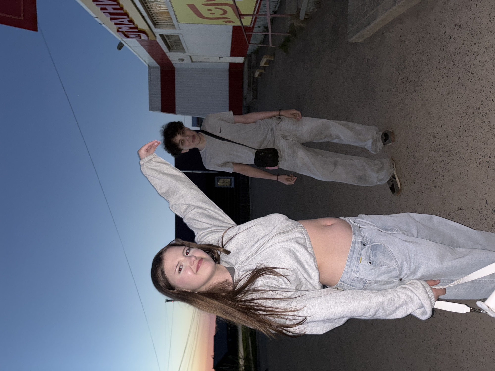 A photograph taken at an angle shows two young individuals in what appears to be an urban setting. The foreground features a female with long brown hair, wearing a cropped gray hoodie and light blue jeans, posing with her arm raised. Her midriff is exposed. Behind her, a young male is visible wearing a light gray t-shirt, matching pants, and a black crossbody bag. They are standing on a paved surface. In the background, there's a building with a red and white facade, part of which is blurred. The sky transitions from blue to a sunset gradient. The location is likely in the city of Фоки, Russia.
