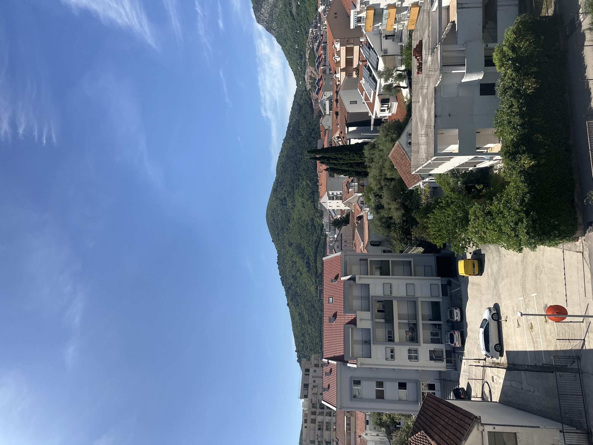 The image presents a bright, sunlit view of a residential area nestled against a large, verdant mountain under a clear blue sky. The sky, occupying the upper and left portions of the frame, is largely cloudless save for a few wispy, high-altitude white clouds.
The left side of the scene is dominated by a steep, green mountain densely covered in trees, suggesting a lush, natural environment. At the base of this mountain and extending into the mid-ground and foreground are numerous multi-story residential buildings. These buildings typically feature white or light-colored facades, many with red-tiled roofs. Balconies are common, and some roofs are equipped with solar panels. Air conditioning units are visible on several building exteriors.
Trees and green hedges are interspersed throughout the urban landscape, adding splashes of natural color among the structures. In the lower right section, what appears to be an asphalt parking lot or street is visible, with several cars parked, including a distinct yellow car, a white car, and a darker vehicle. An orange basketball backboard stands prominently in this area.
The time of day is bright midday or early afternoon, indicated by the strong, direct sunlight. The weather is clear and sunny. The architectural style and natural setting are consistent with a Mediterranean or Adriatic coastal town, aligning with the given location context of Budva, Montenegro. No specific human activity is captured, and no legible text is visible in the frame.