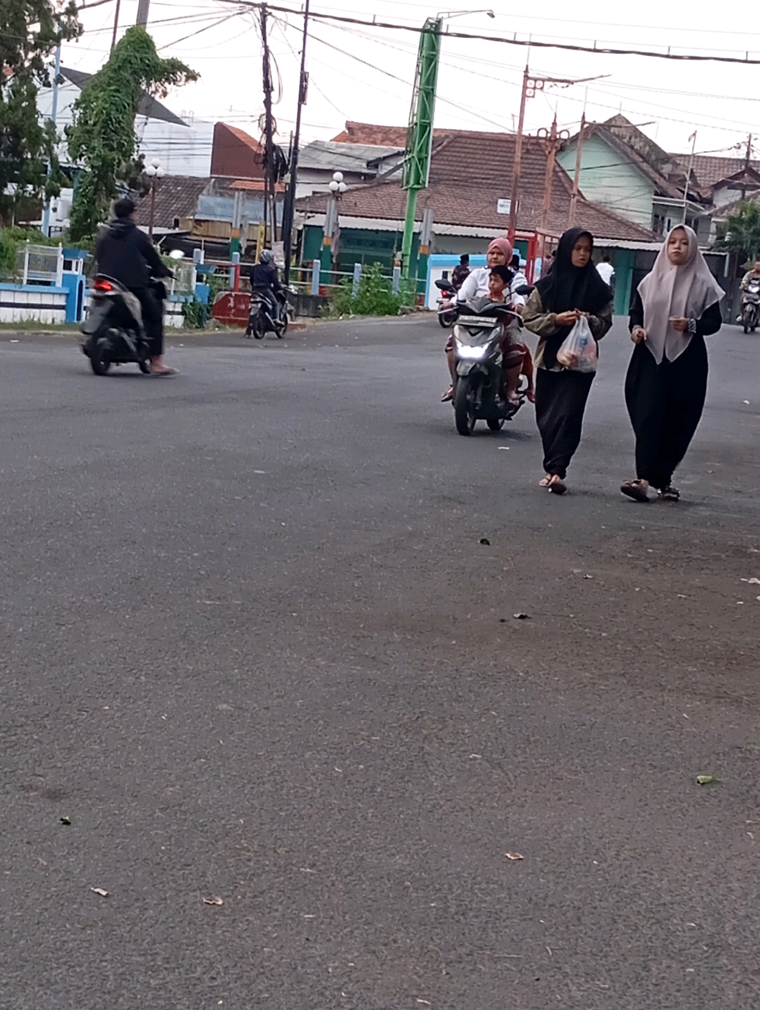 In Kludan, Indonesia, a cloudy day sees two women walking on an asphalt road, their bodies covered in dark clothing and headscarves. One woman, on the left, wears a light grey headscarf and a black garment, carrying a clear plastic bag filled with items. The other woman, to her right, is dressed in black, her headscarf also black, and she carries nothing. Behind them, a young child sits on a scooter with a woman in a pink headscarf. Further in the background, a man on a scooter rides away from the viewer, his back turned. The scene is bordered by a low blue and white fence and buildings with tiled roofs.