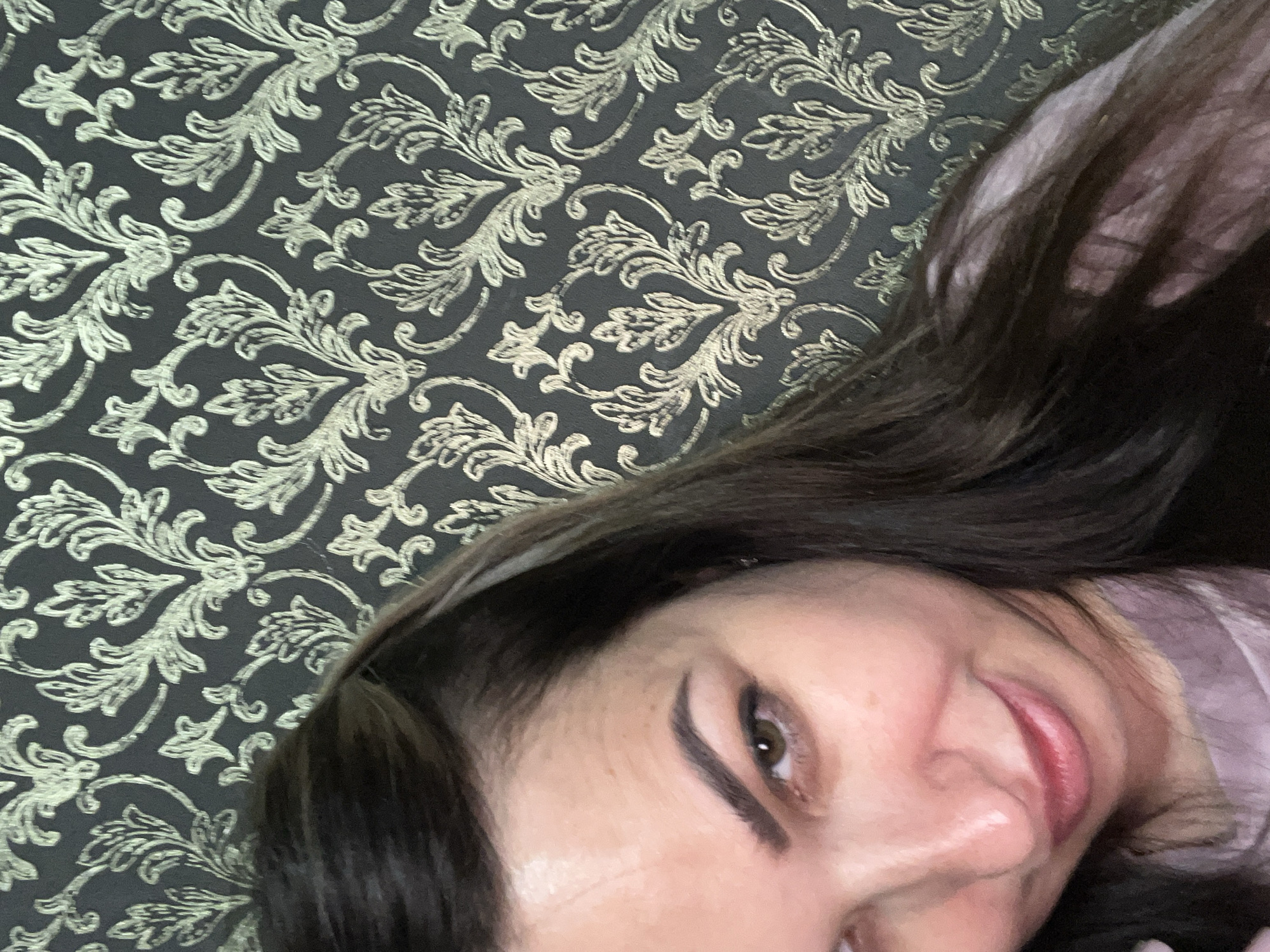 A close-up, high-angle selfie shows the upper body of a woman with long, dark brown hair. She is looking downwards and to her right with a slight smile. Her left eye, eyebrow, and the bridge of her nose are visible. She is wearing a light purple shirt. The background is a dark grey wallpaper with a repeating pattern of gold floral motifs. The scene appears to be indoors.