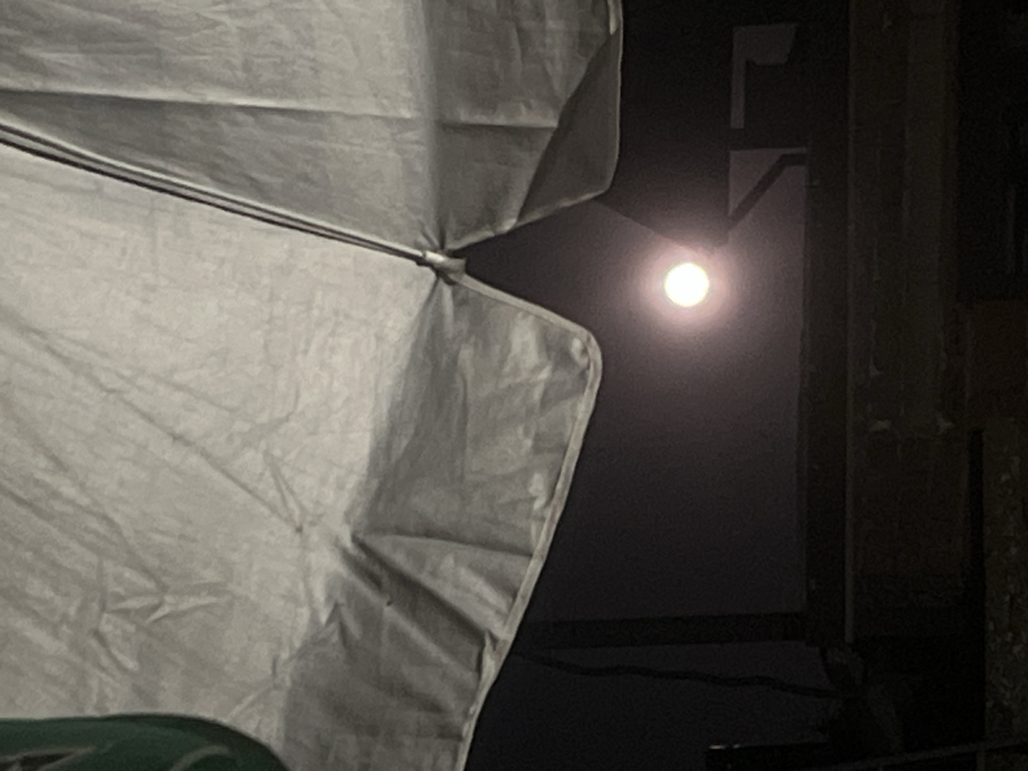 The image shows a partially obscured view of a bright, full moon against a dark sky. The moon is positioned centrally in the lower portion of the frame, emitting a soft glow. Above the moon, the underside of a large, light-colored umbrella or canopy is visible, its fabric creased and textured. Part of a dark, shadowy structure, possibly a building or scaffolding, is visible behind the moon. The foreground is dark and out of focus, showing hints of what might be textured surfaces. The scene is likely an outdoor setting at night, with the moon being the primary light source. The context provided about Belashtitsa, Bulgaria, suggests this might be a view from a residential or public outdoor space within that location.