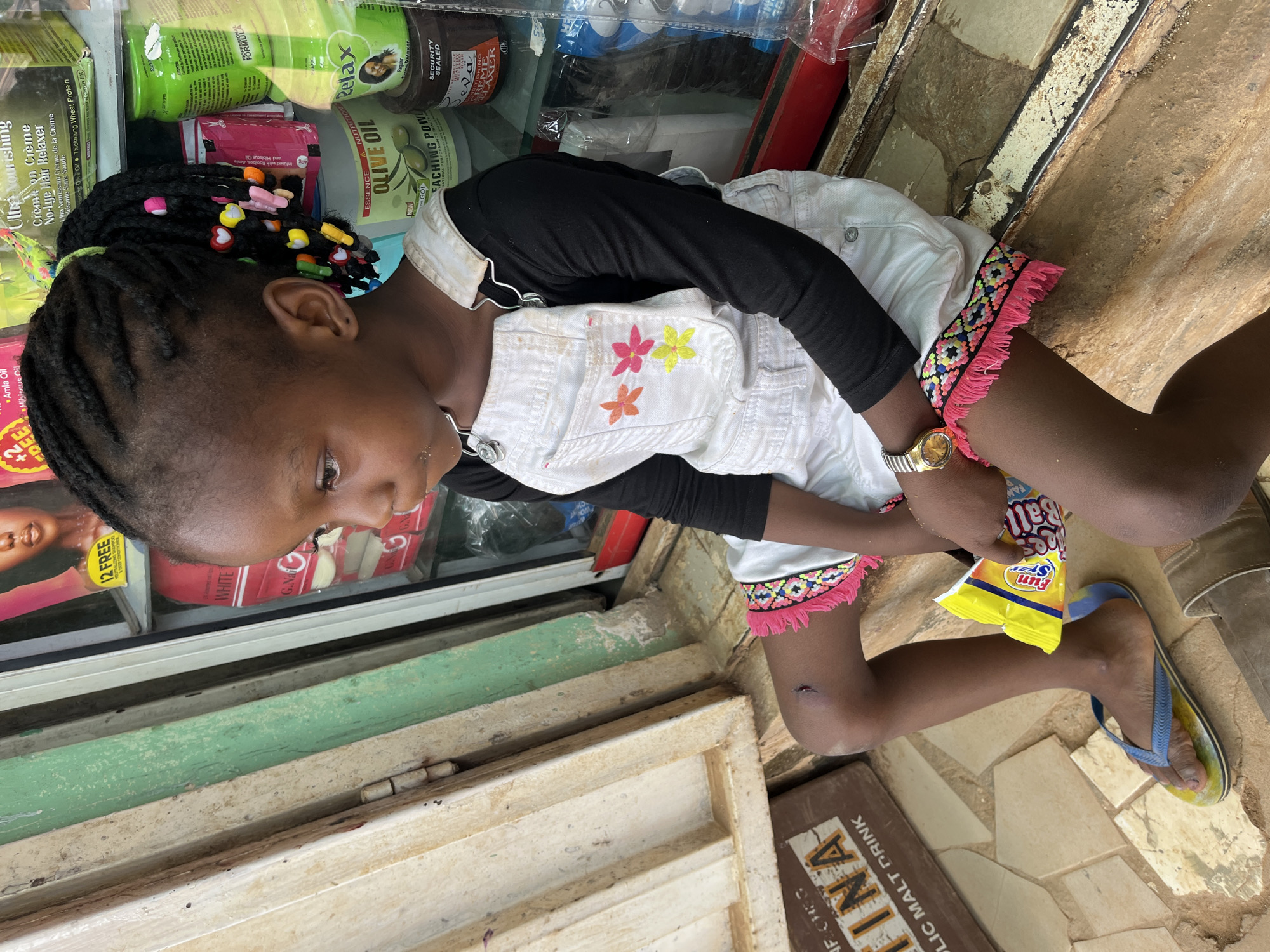 A young girl is crouching outside a shop in what appears to be Madalla, Nigeria. She is wearing a white overall dress with floral decorations, a black long-sleeved shirt underneath, and blue and yellow flip-flops. Her hair is braided with colorful beads. She holds a bag of "Fun Size Ball" candy. The shop behind her displays hair relaxer products and other items. The ground is tiled, and a sign reading "INA MALTC DRINK" is visible.
