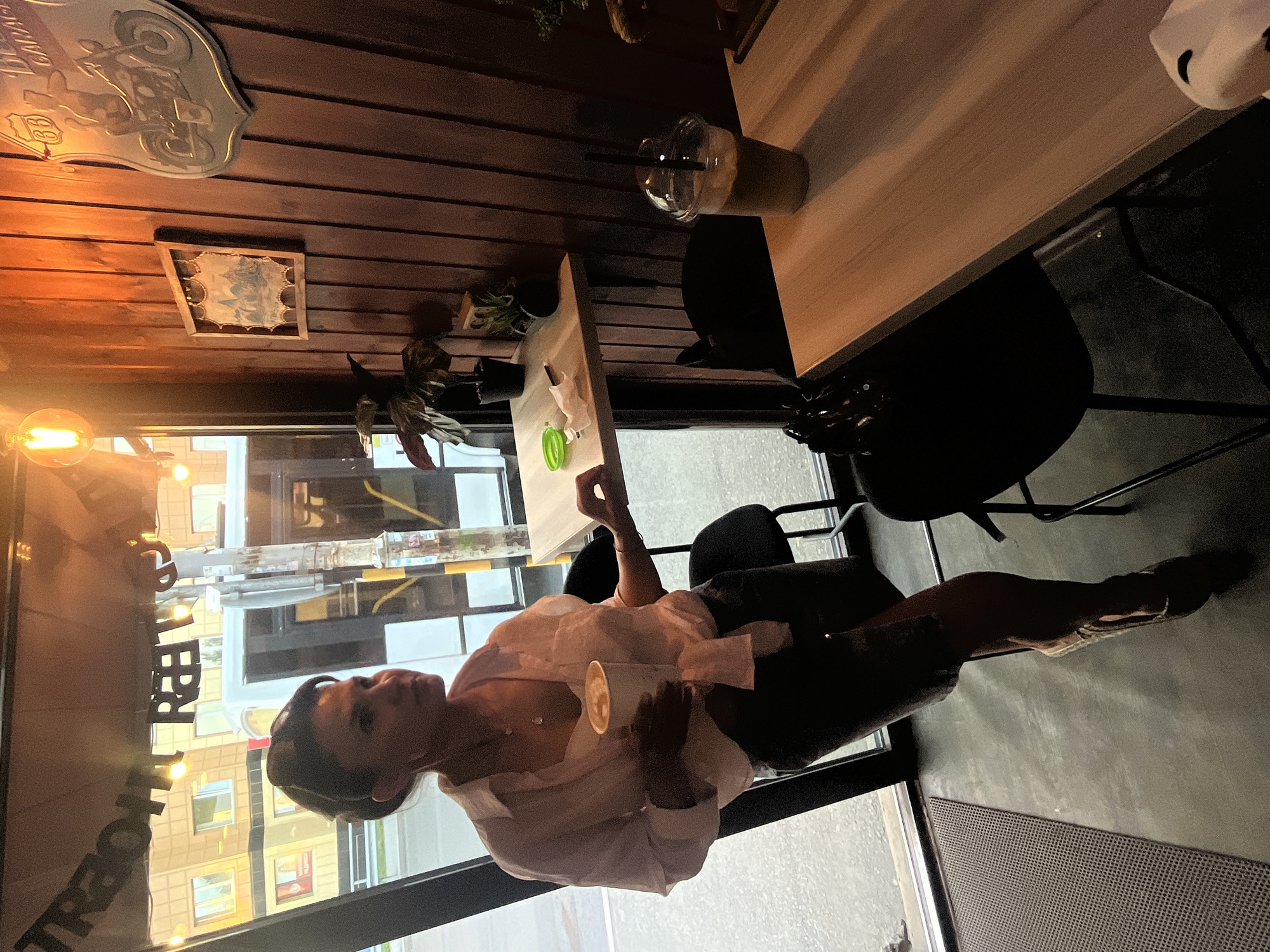 A woman stands in a cafe, holding a cup of coffee and looking to the left. She is wearing a light pink unbuttoned shirt, a white top, and dark shorts. The cafe has wooden walls and is decorated with framed pictures and a motorcycle-themed sign. There are two tables with chairs. Outside the window, a bus and buildings are visible, suggesting an urban setting. The lighting in the cafe is warm and atmospheric, with a visible light bulb hanging from the ceiling. The overall impression is that of a casual, comfortable cafe environment.