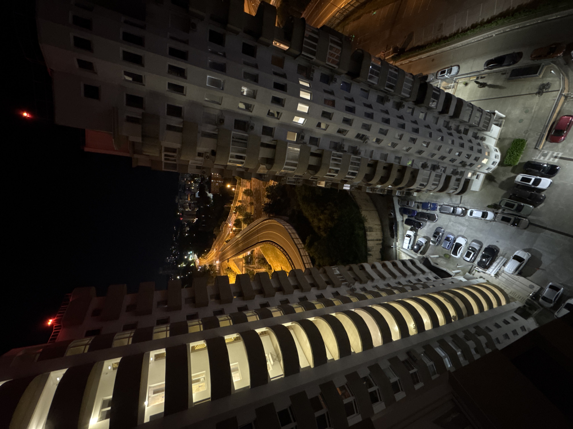 An aerial, high-angle nighttime shot shows two tall, curved apartment buildings with balconies, illuminated from within. Red lights are visible atop one of the buildings. A highway curves through a wooded area between the buildings, with streetlights and car headlights visible. Below, a parking lot is filled with cars. The scene is identified as Sochi, Russia.