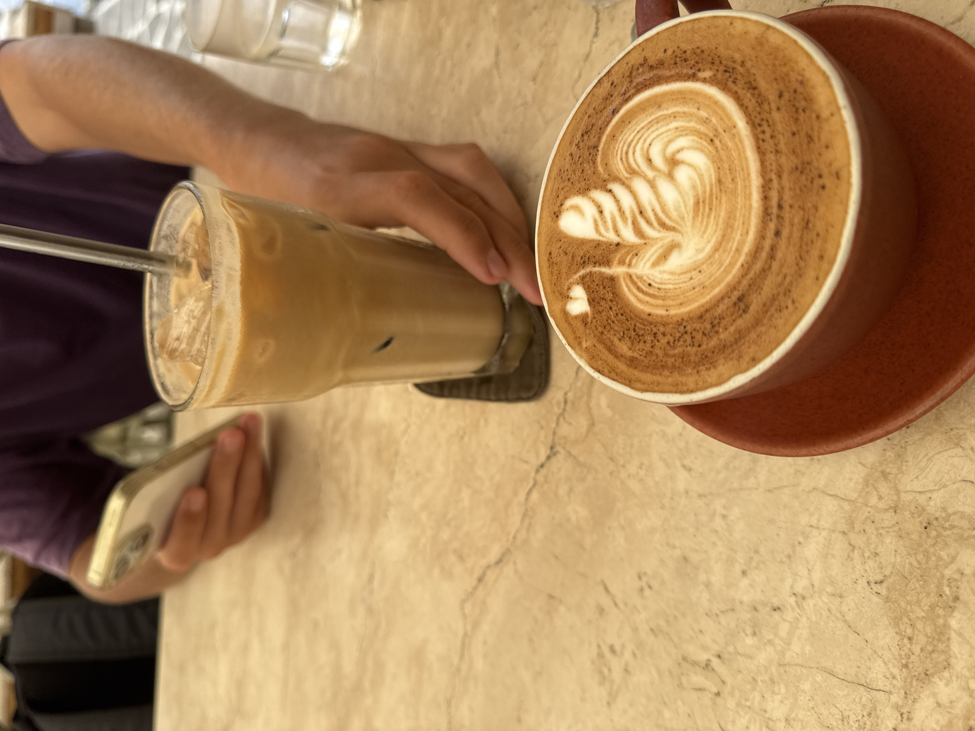 A top-down, slightly angled shot captures a cafe table with two coffee beverages and parts of a person's hands and arm. On the left, a tall glass filled with iced coffee, ice cubes, and a metal straw rests on a dark coaster. The iced coffee is a light, creamy brown color. To its right, a ceramic cup of hot coffee, likely a latte or cappuccino, sits on a red-brown saucer. The hot coffee features intricate white latte art, resembling a fern or wing pattern, against a rich brown background, with tiny dark specks around the edge of the foam.
A person's arm, dressed in a purple short-sleeved top, extends from the upper left, with one hand positioned near the iced coffee. A second hand is visible lower down, holding a light-colored smartphone. The table surface is a light beige or cream-colored material, possibly stone or marble, characterized by subtle veining and a slightly speckled texture. A clear glass is partially visible in the upper background. The bright lighting suggests it is daytime. The scene conveys a casual, relaxed atmosphere, typical of a cafe visit. No discernible text is visible.