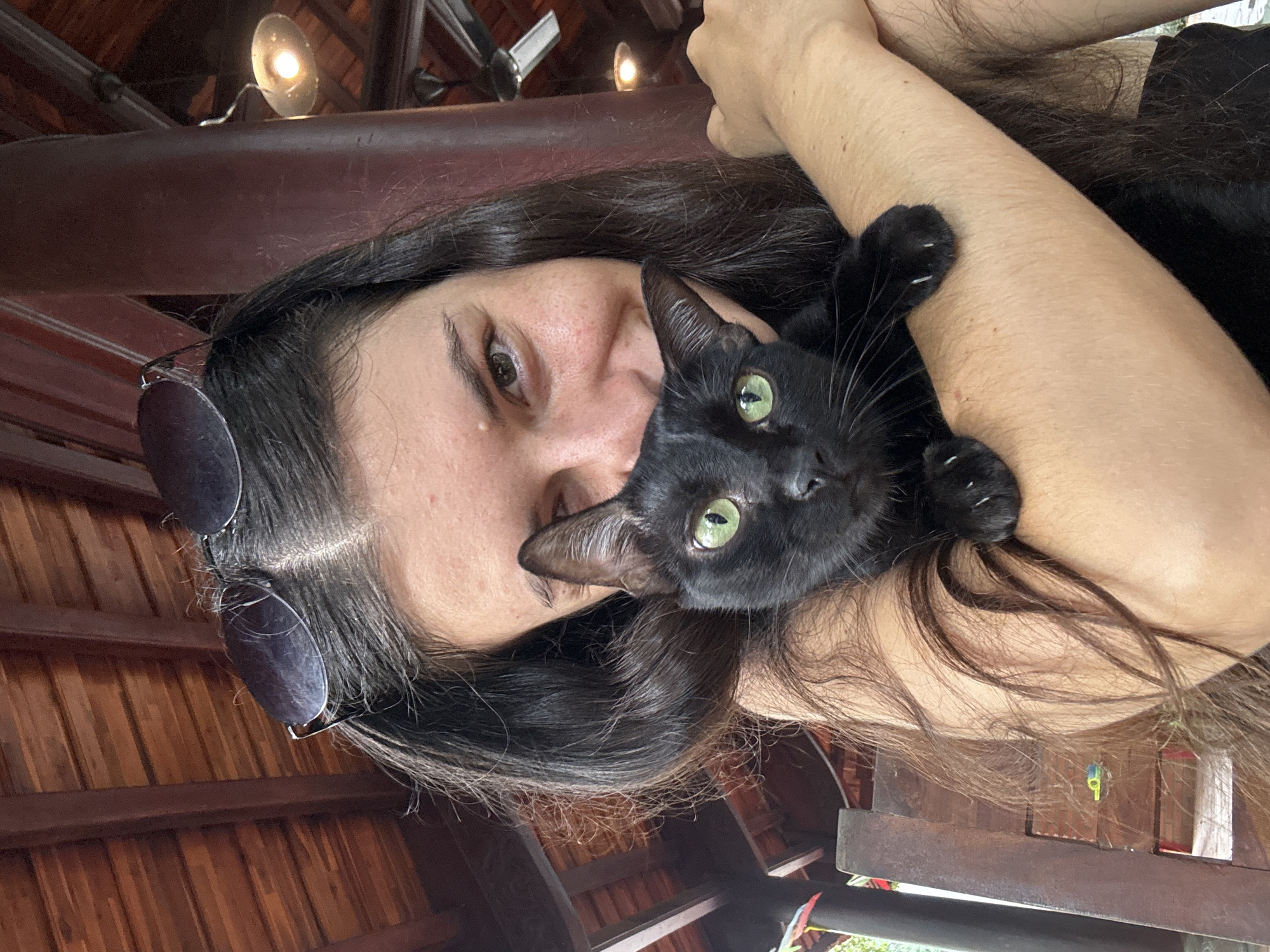 In Mũi Né, Vietnam, a woman with long, dark hair and sunglasses perched on her head is holding a black cat. The cat has striking green eyes and is nestled in her arms. They are outdoors, with a wooden ceiling and a lamplit structure visible in the background, suggesting a covered seating area. The woman is smiling and looking at the camera, while the cat is also looking directly at the viewer. A wooden chair is partially visible to the left of the frame.