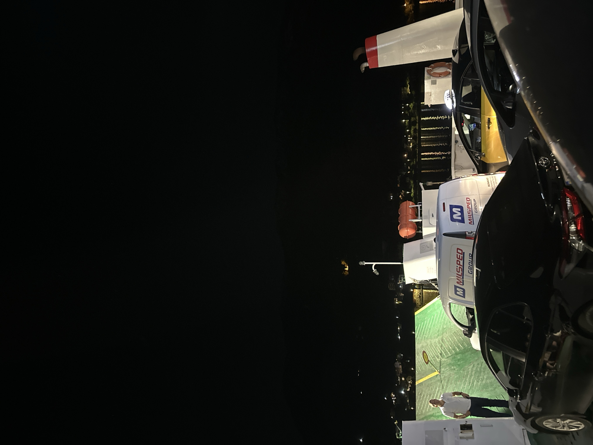 The image, taken at night, shows the deck of a ferry in Lepetane, Montenegro. A man in a white shirt and dark pants stands on the green painted deck near a white Milsped Group van and a dark car. Other cars and a yellow taxi are visible. In the background, lights from buildings on the shore reflect on the water. A lifeboat is visible near the ferry's structure.