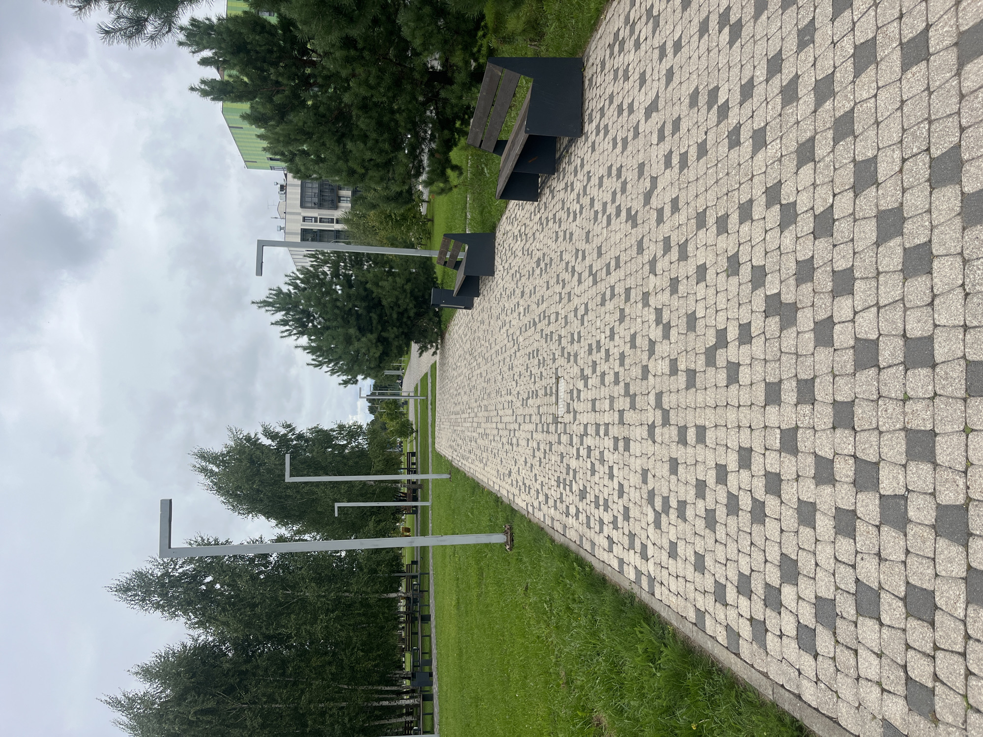 The image depicts a park pathway in Innopolis, Russia, with a cloudy sky overhead. The pathway is paved with light and dark grey interlocking stones, creating a checkerboard pattern. To the left of the pathway is a well-maintained lawn bordered by trees. Modern, minimalist lampposts with L-shaped fixtures are spaced along the left side of the pathway. On the right side of the pathway, there are two dark grey benches with wooden slats for seating, situated amongst evergreen trees. In the background, contemporary apartment buildings with green and white facades are visible. The scene is clean and orderly, suggesting a developed urban park area. There are no people visible in the image.
