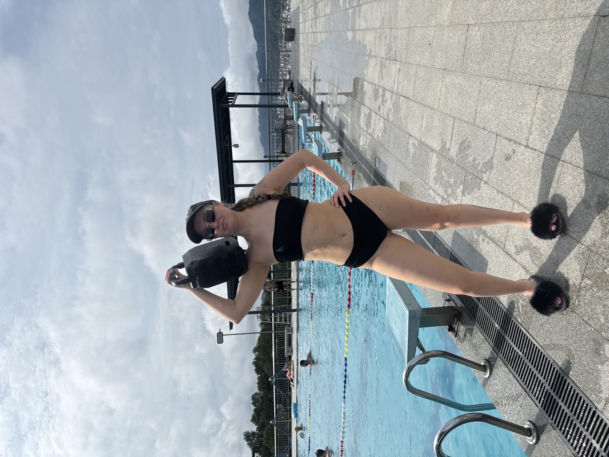 A woman in a black bikini and furry slides stands by a swimming pool in Yiwu, China. She is wearing a baseball cap and sunglasses and has a speaker on her right shoulder. The pool is blue and has lane markers. There are other people in the pool and in the background, along with trees and buildings. The sky is cloudy.