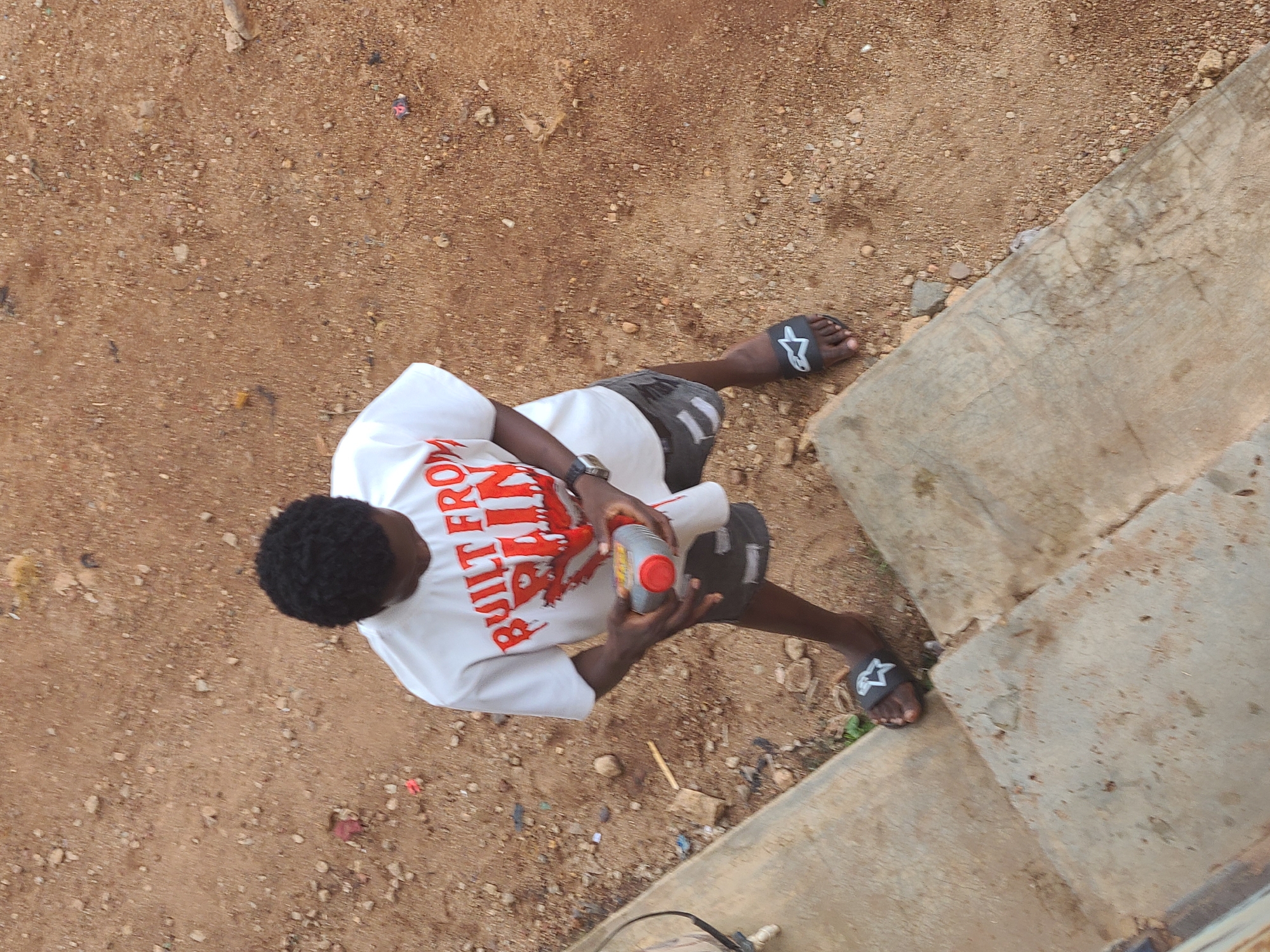 A medium, slightly downward-angled shot reveals a Black individual wearing a white t-shirt with the phrase "BUILT FROM PAIN" in red, with black and gray shorts, and black flip-flops with white stars on them. The person is holding a gray and red plastic bottle with both hands. They are standing next to a concrete staircase. The ground is covered in brown dirt and gravel, with small rocks scattered around. The location is Gura Top, Nigeria.