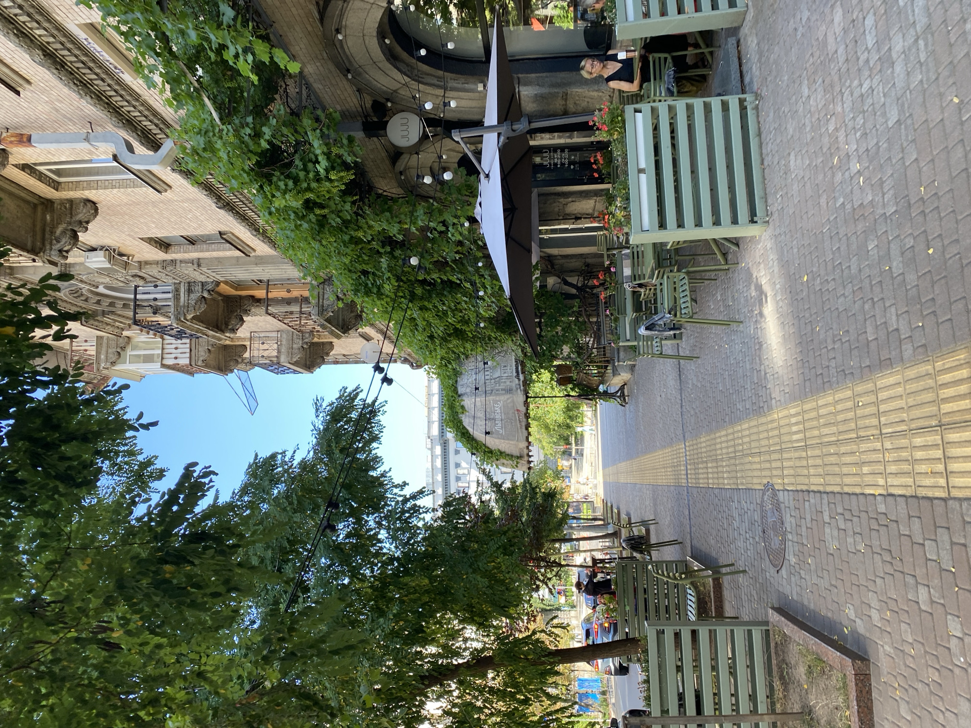 This is a vertical, medium shot of an outdoor cafe on a city street. The cafe has several green metal tables and chairs, some with umbrellas, set up along a brick sidewalk. A woman in a black dress is seated at one of the tables, looking down at a menu. Trees with green leaves line the sidewalk, casting shadows on the ground. The cafe is attached to a building with ornate balconies and a brick facade, covered in ivy. In the background, a street with cars and pedestrians can be seen. The sky is clear and blue.