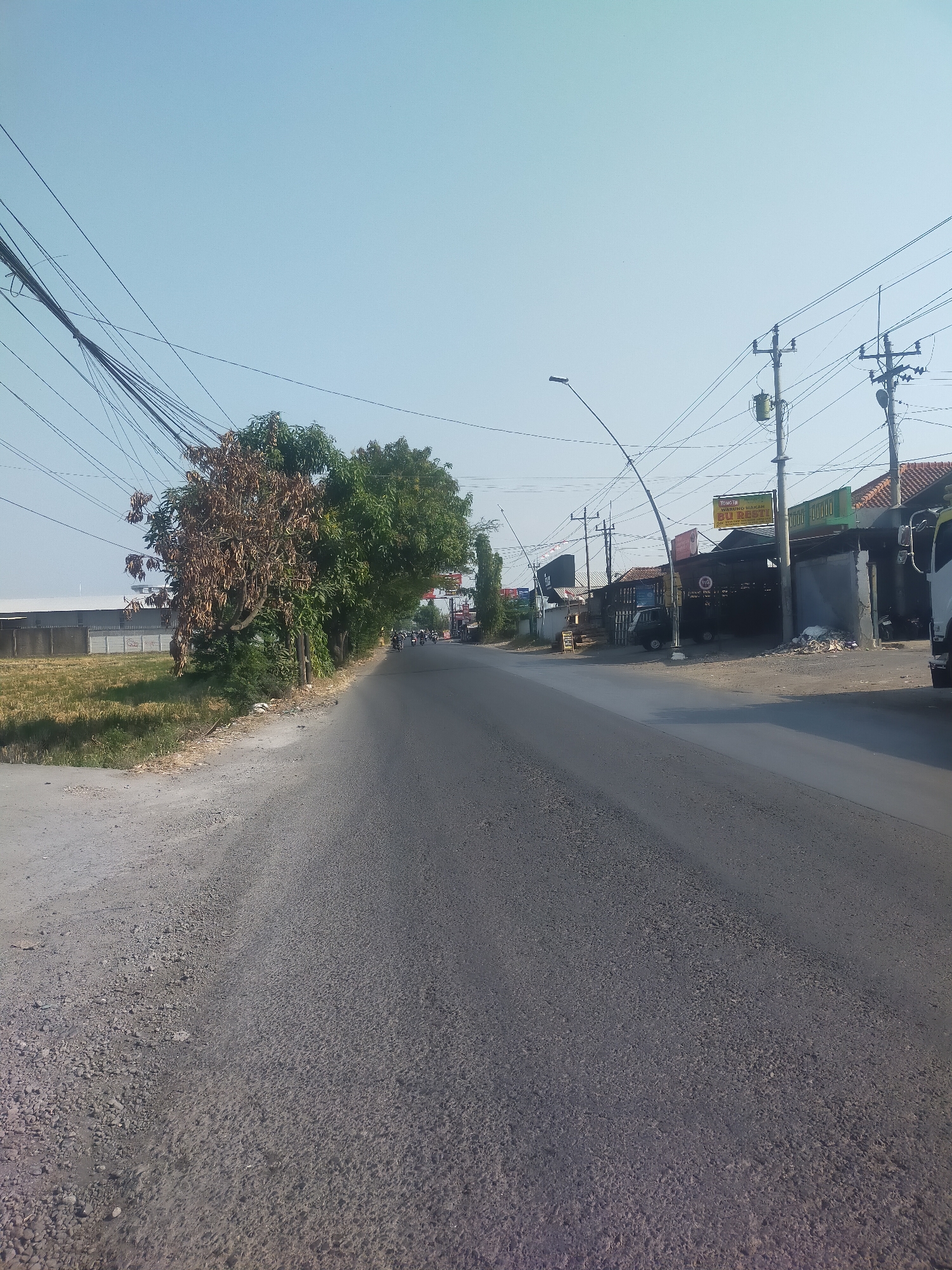 The image depicts a street scene in Pesarean, Indonesia. The road, paved with asphalt and gravel on the shoulder, is surrounded by a mixture of residential and commercial buildings, as well as vegetation. Overhead, a dense network of electrical wires crisscrosses the sky, supported by utility poles. To the left of the road, a field of dry grass is visible, bordered by a long, low building with a light-colored wall. Further down the road, trees line the left side, one of which appears to be dead or dying. On the right side, a row of buildings houses various businesses, indicated by signs such as "TOKO IBUM MAKAN BU RESTU" and others in Indonesian. Several vehicles are present, including a dark-colored car, a white truck, and multiple motorcycles carrying passengers, suggesting ongoing traffic. The sky is clear and bright, indicating daytime.