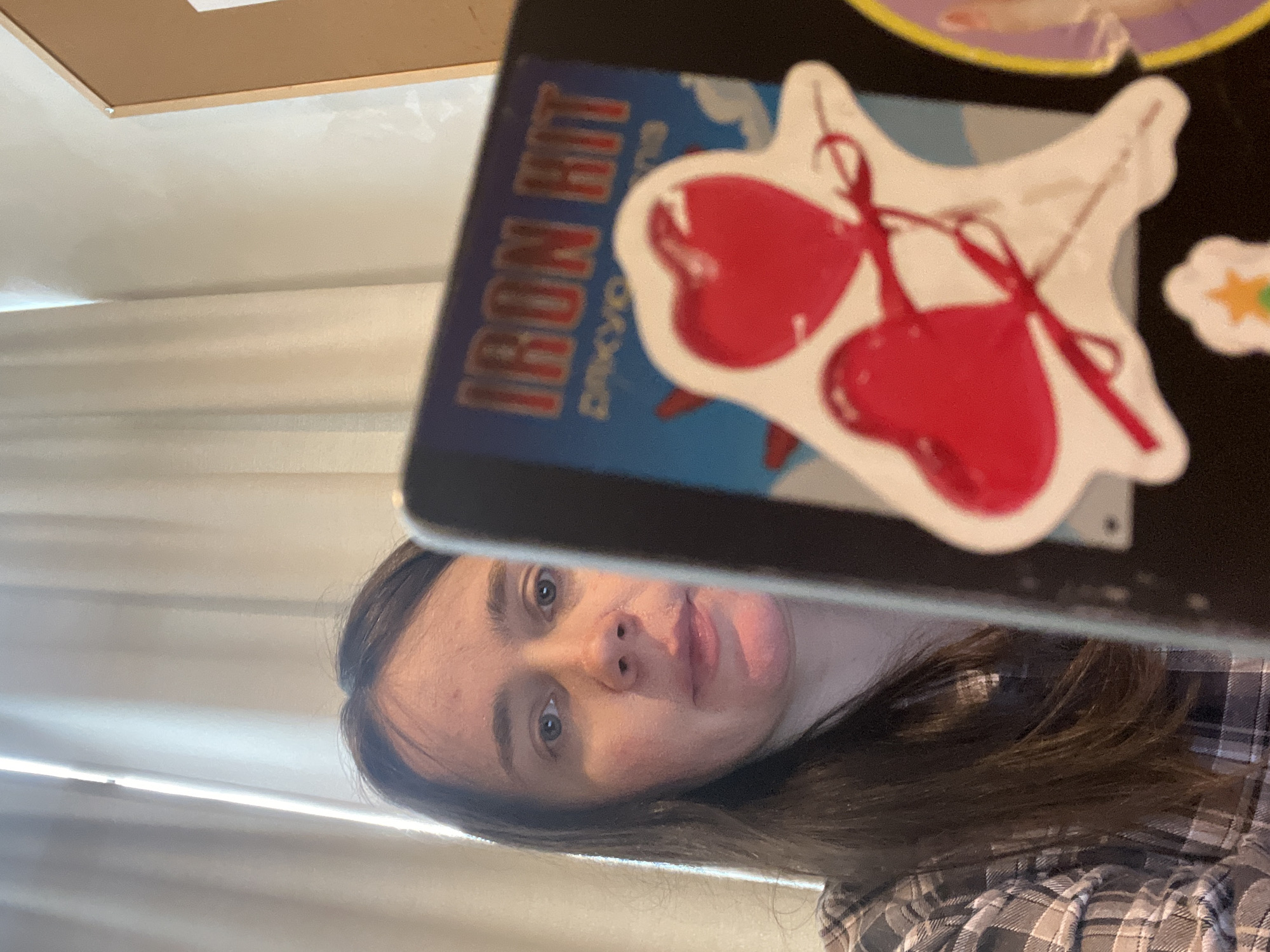 A person with long brown hair, wearing a plaid shirt, is looking at the camera. They are holding a tablet in front of their face, obscuring part of it. The tablet has stickers on it, including two red hearts tied with red ribbon and a sticker with the words "IRON HIT" in red lettering. The background appears to be a room with light-colored curtains. The scene is lit by natural light. There is no information to suggest this image was taken in Kyiv, Ukraine.