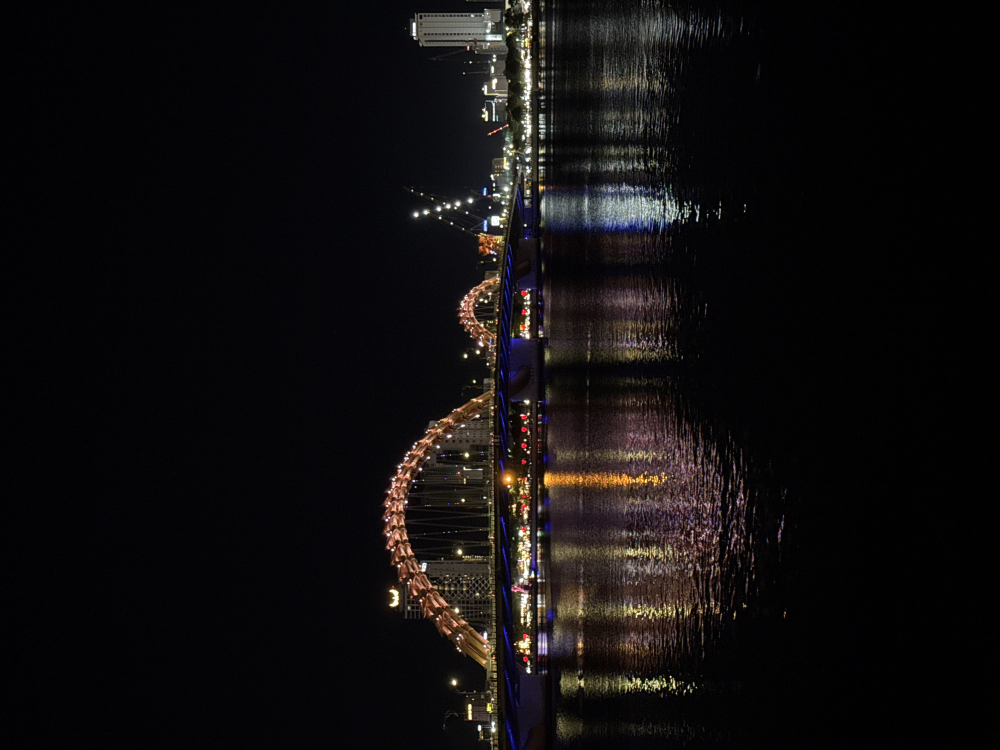 The image displays a nighttime view of the Han River in Đà Nẵng, Vietnam, featuring the illuminated Dragon Bridge (Cầu Rồng). The bridge's prominent dragon-shaped arch is illuminated with golden-orange lights, spanning horizontally across the river. The bridge deck below the arch also features various artificial lights, primarily blue and white. On both banks of the river, the city's urban landscape is visible, characterized by illuminated multi-story buildings and distant high-rise structures. Several construction cranes with lights are discernible on the far right bank. The dark surface of the Han River reflects the bright lights from the bridge and the cityscape, creating elongated, shimmering patterns of orange, blue, and white. The sky above is uniformly black. No individuals or discernible actions are present within the frame. The scene is static, showcasing the illuminated urban infrastructure.