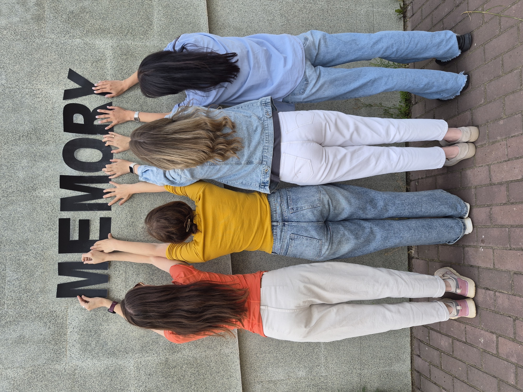 Four individuals are captured from an overhead perspective, lying prone on a grey, textured surface, appearing to spell out a word with large black block letters. Their backs are to the viewer, and they are arranged vertically, with their heads near the top of the image and their feet towards the bottom.
The word formed by the arrangement of the people and the letters is "MEMORY". Each person is positioned next to or interacting with parts of the letters.
From top to bottom:
The first person has long dark hair and is wearing a light blue long-sleeved shirt and light blue jeans. Their hands are placed on the letter 'Y'.
The second person has wavy blonde or light brown hair and is dressed in a light blue denim jacket over a white top and white pants. They wear a watch on their left wrist and are holding onto the letter 'R'.
The third person has brown hair, possibly tied back, and is wearing a yellow long-sleeved shirt and blue jeans. Their hands are on the letter 'O'.
The fourth person has long dark brown hair and is wearing an orange long-sleeved shirt and light beige or off-white pants. A smartwatch is visible on their left wrist, and they are touching the letter 'M'.
The surface they are on transitions to a red brick pavement on the right side of the image, with some green foliage visible near the edge. The lighting suggests it is daytime, possibly outdoors.