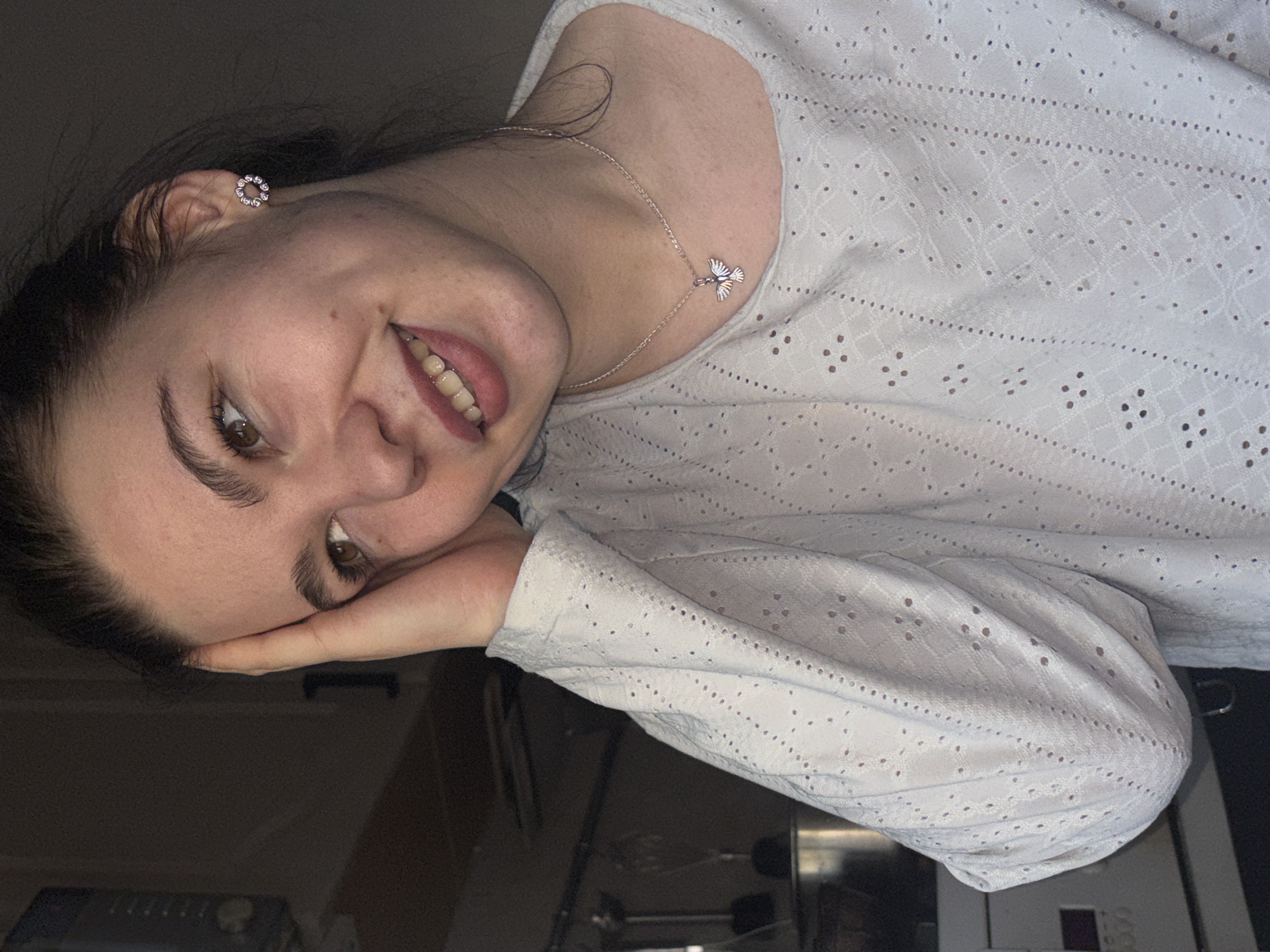 A young woman with brown hair and eyes smiles at the camera. She is wearing a white, eyelet-patterned long-sleeved shirt and a silver necklace with a bird-shaped pendant. A silver, circular earring is visible in her left ear. Her left hand is resting on her face. The background is dark and indistinct, but hints of furniture and appliances suggest an interior setting, possibly a home or apartment in Perm, Russia.