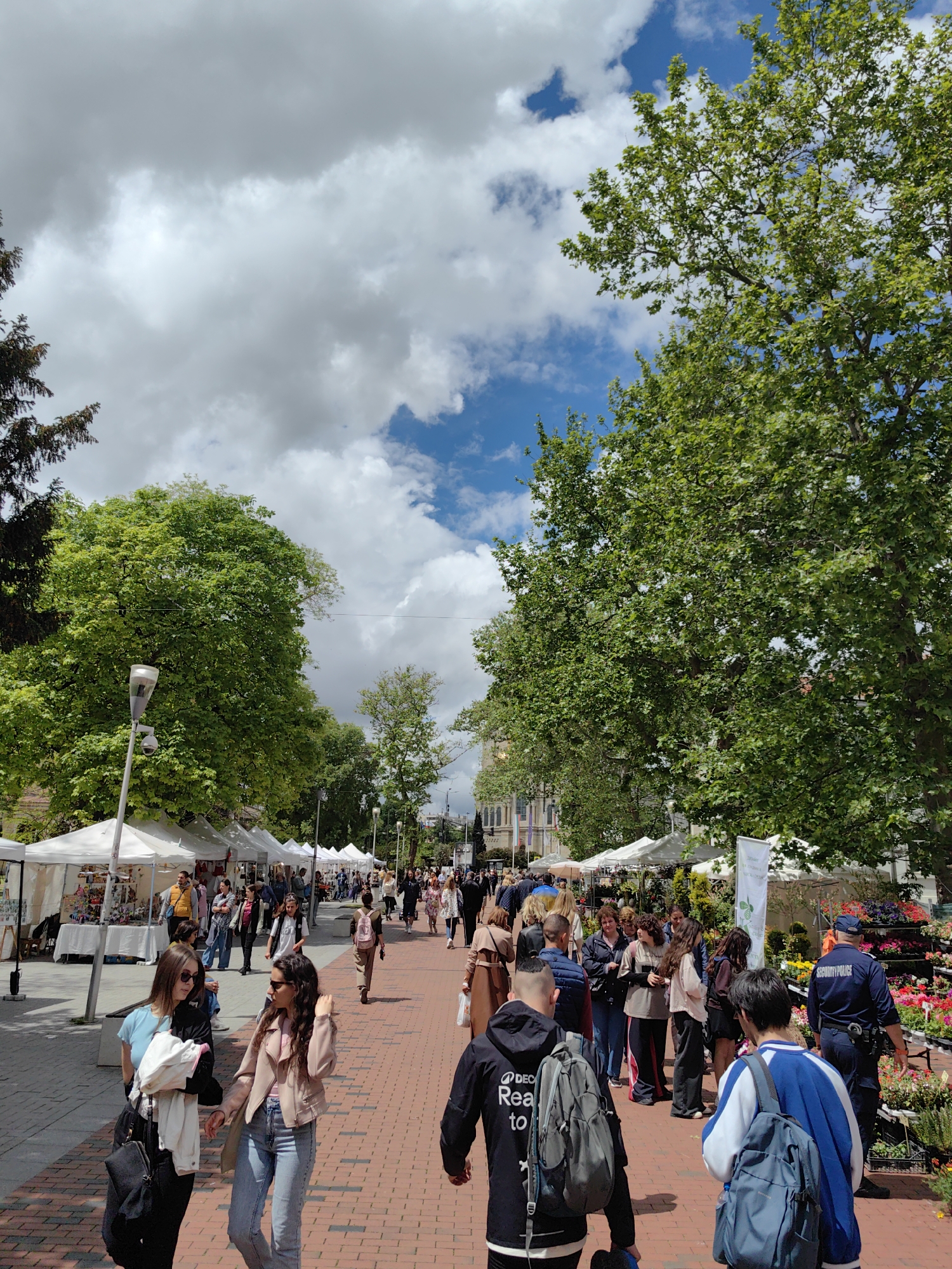 The image shows a bustling pedestrian walkway in Varna, Bulgaria, populated with people, trees, and vendor stalls. The walkway is paved with red bricks. People are walking in both directions, some in small groups, others alone. Several white tents are lined up along the walkway, likely vendor stalls. A "SECURITY POLICE" officer is present. On the right side of the image is a display of flowers. The sky is filled with white clouds and patches of blue. Trees frame the scene on both sides. A lamppost is visible.